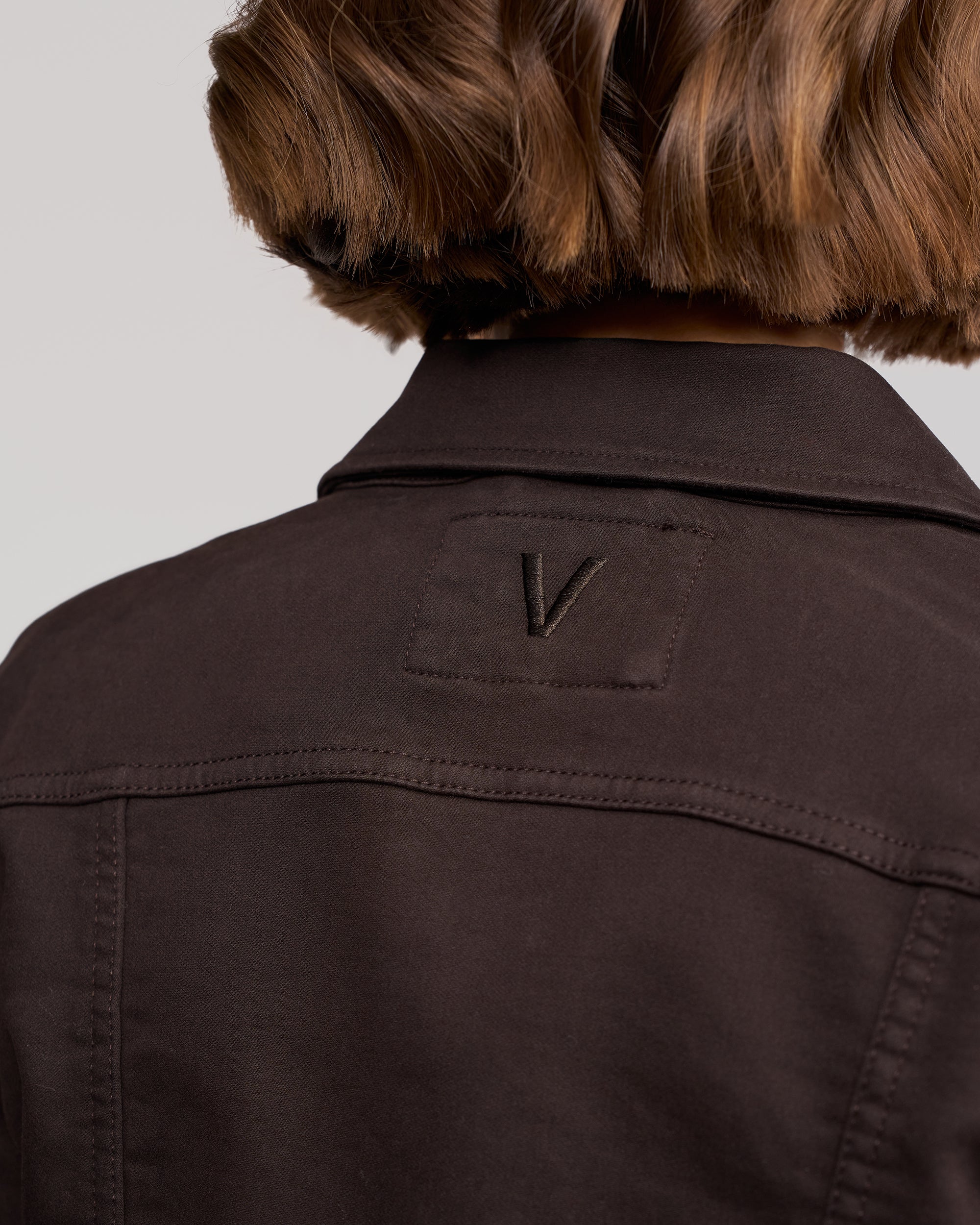 A close-up shows the upper back of a person wearing the Vassalli Rider Knit Jacket in chocolate, featuring a stitched 'V' patch. The relaxed fit complements their short, wavy brown hair.
