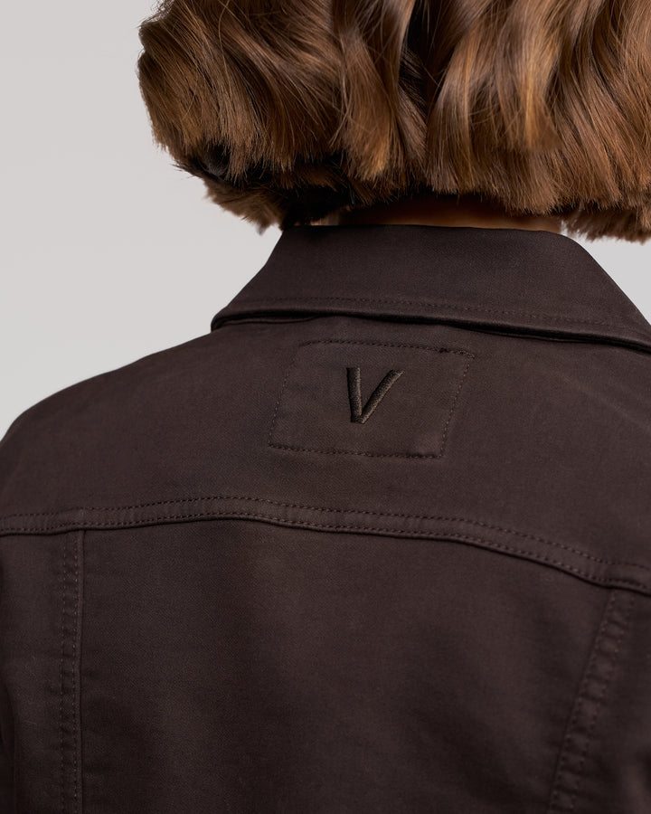 A close-up shows the upper back of a person wearing the Vassalli Rider Knit Jacket in chocolate, featuring a stitched 'V' patch. The relaxed fit complements their short, wavy brown hair.
