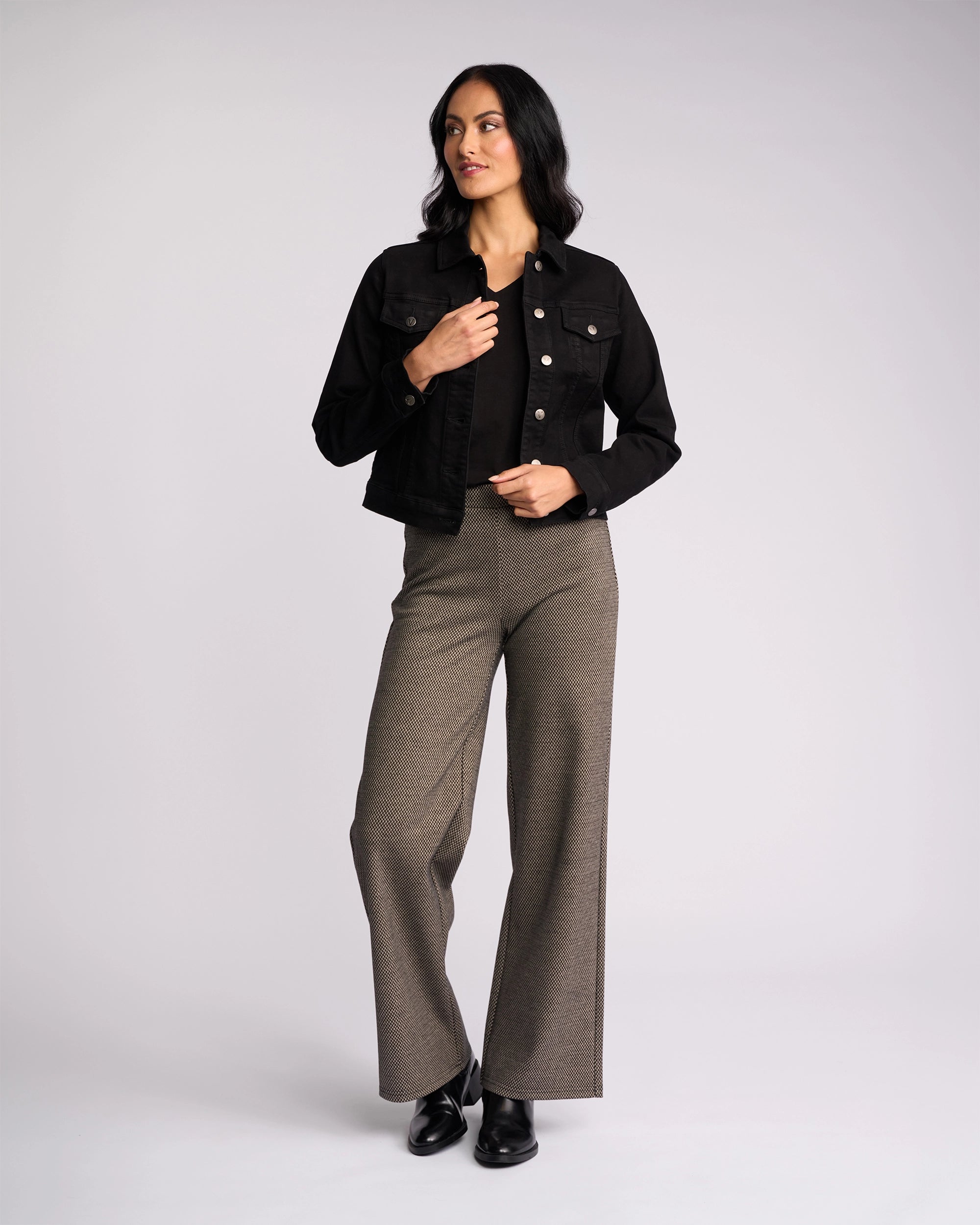 A person stands against a plain background wearing the Vassalli Rider Knit Jacket in black, paired with grey high-waisted wide-leg pants and black shoes. They have long dark hair and look slightly to the side, one hand touching the jacket.