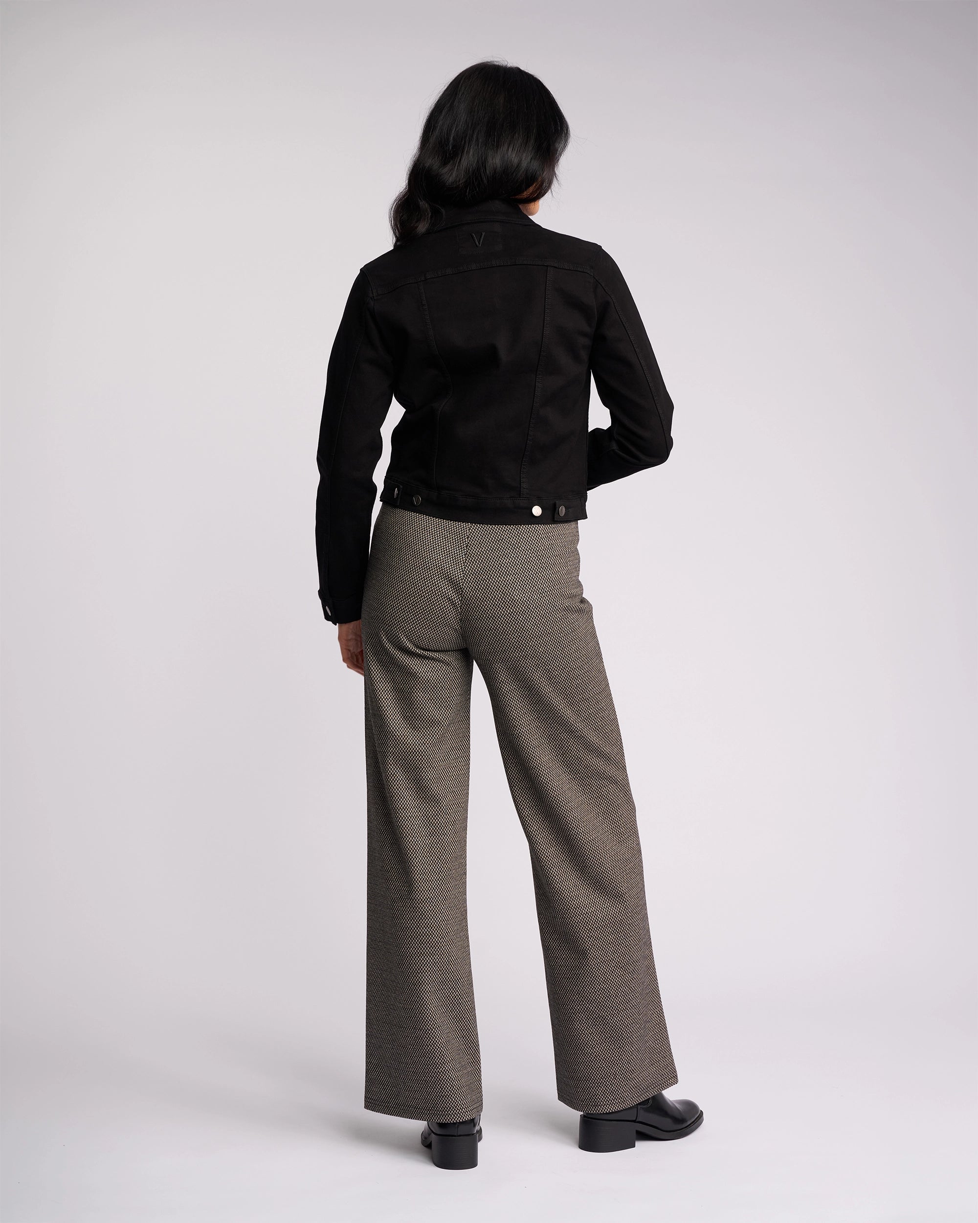 A person with long dark hair faces away, wearing the Vassalli Rider Knit Jacket in black, gray wide-leg trousers, and black shoes against a plain light background.