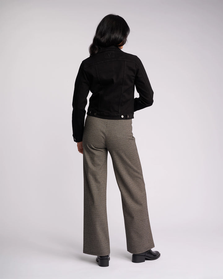 A person with long dark hair faces away, wearing the Vassalli Rider Knit Jacket in black, gray wide-leg trousers, and black shoes against a plain light background.