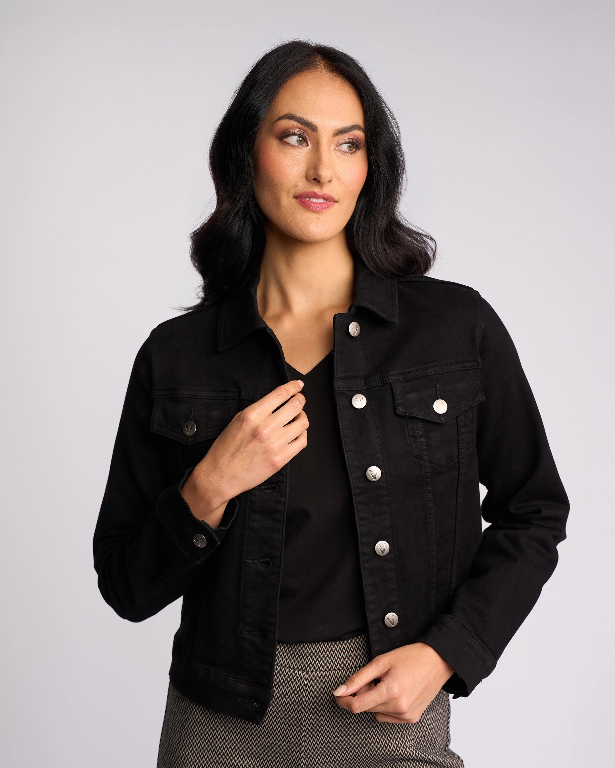 A woman with long dark hair wears the Vassalli Rider Knit Jacket in black over a black top and patterned pants. She stands against a plain light background, looking slightly to the side with a soft smile.