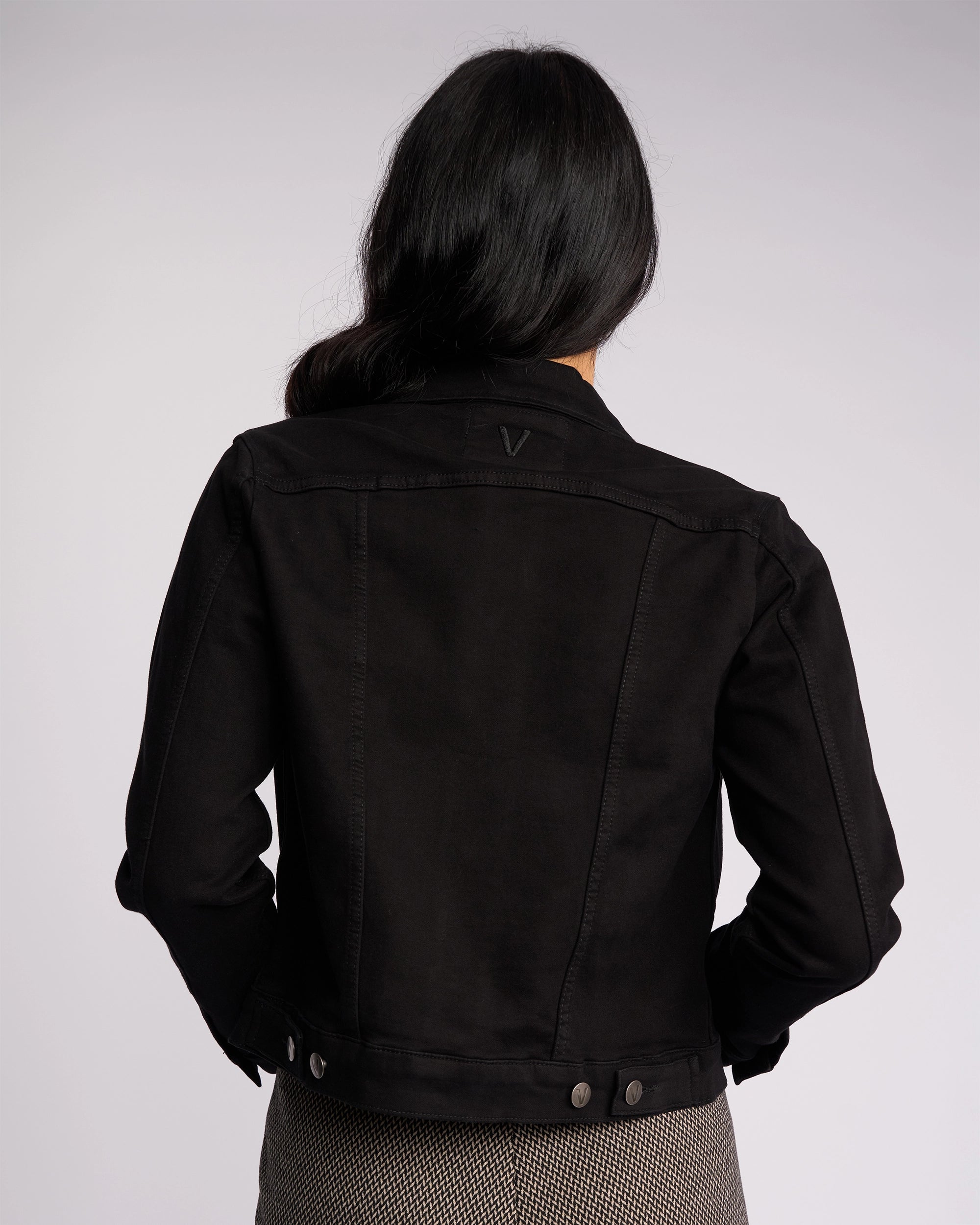 A person with long dark hair, seen from behind, wears the Vassalli Rider Knit Jacket in black and patterned pants against a plain light background.