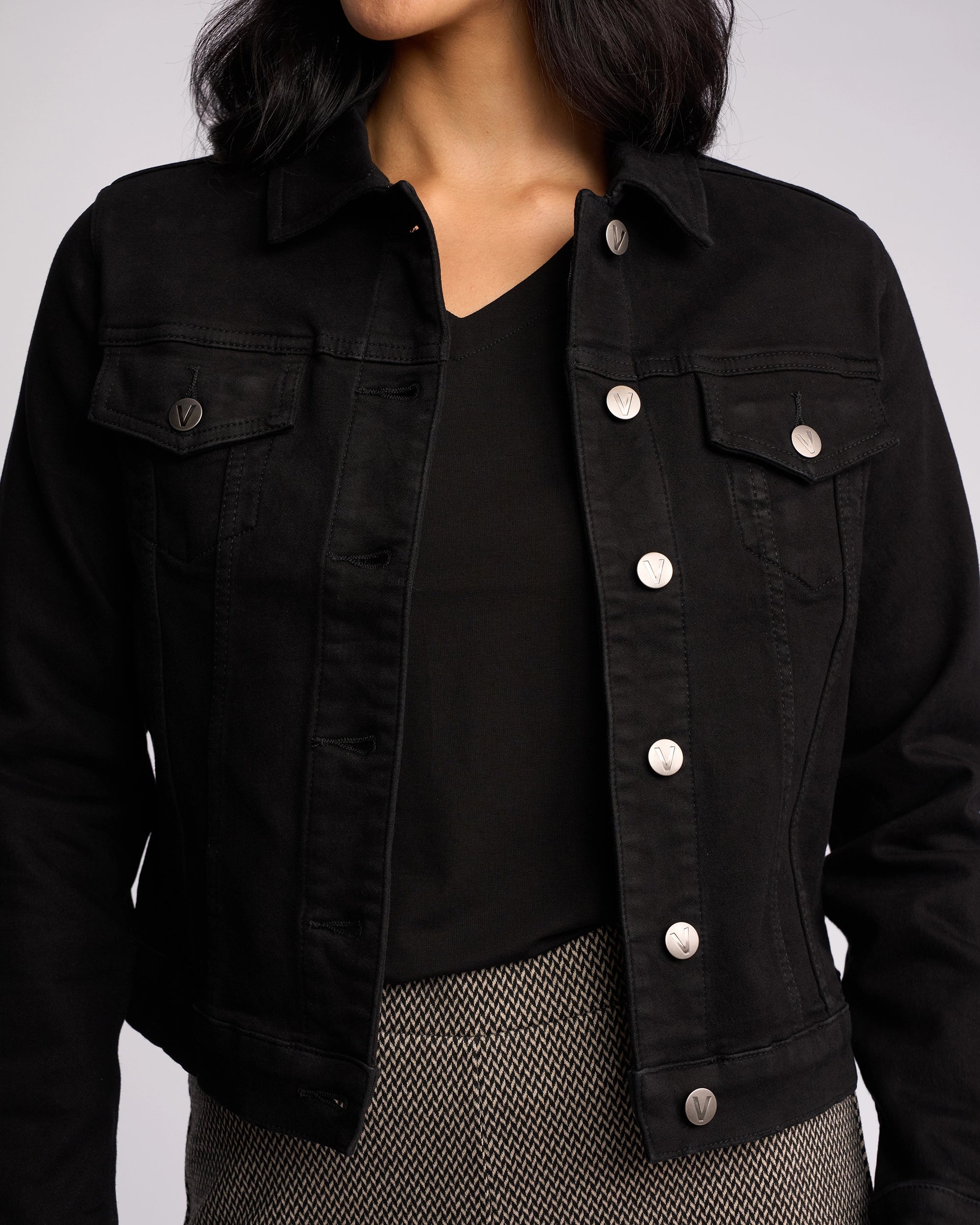 A woman wears the Vassalli Rider Knit Jacket in Black, featuring silver buttons and a relaxed fit, over a black top and patterned pants. Her wavy dark hair is visible, but her face is partially out of frame.