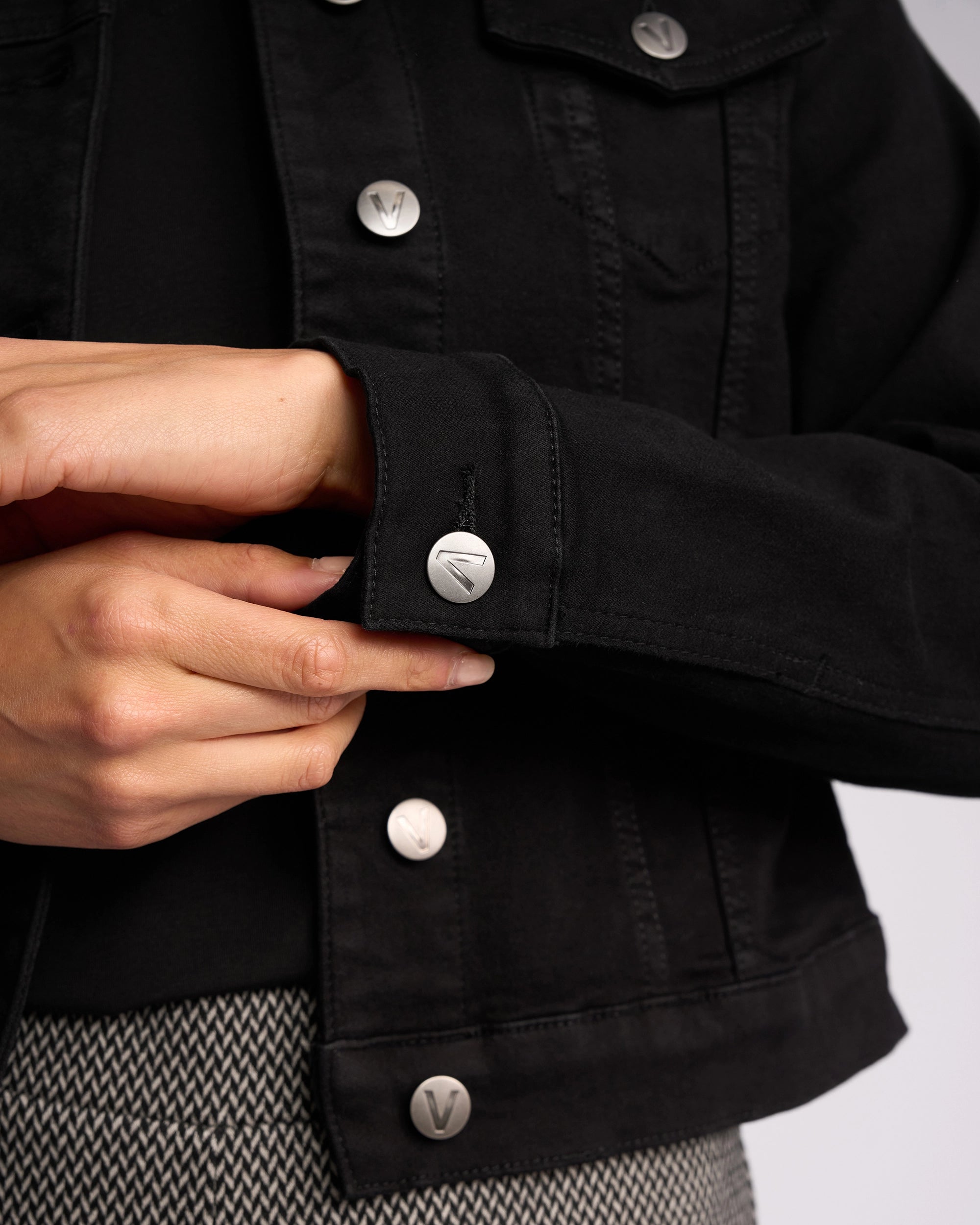 A person fastens the cuff button on the sleeve of a Vassalli Rider Knit Jacket - Black, featuring silver buttons and a relaxed fit. The jacket pairs well with patterned pants. Only the hands and torso are shown.