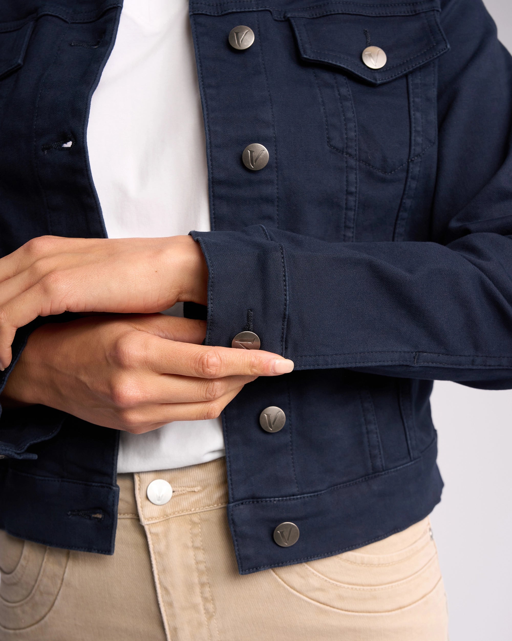 A person, visible from torso to hands, fastens the cuff of a Vassalli Rider Knit Jacket - Carbon, layered over a white shirt and beige pants.