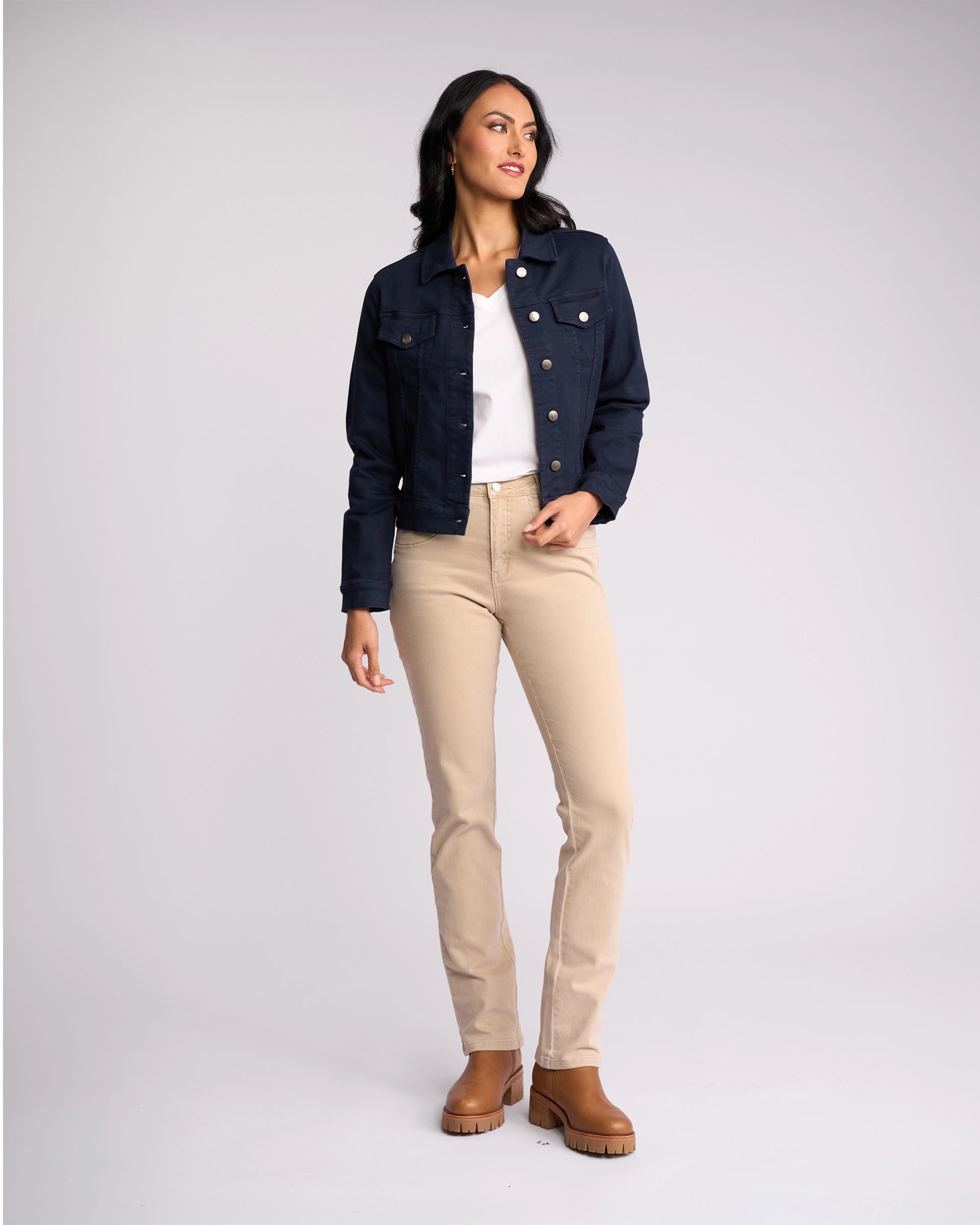A woman stands against a plain background wearing the Vassalli Rider Knit Jacket - Carbon over a white shirt, beige pants, and brown ankle boots. She looks to the side with one hand in her pocket, showcasing the jacket’s stretch comfort.
