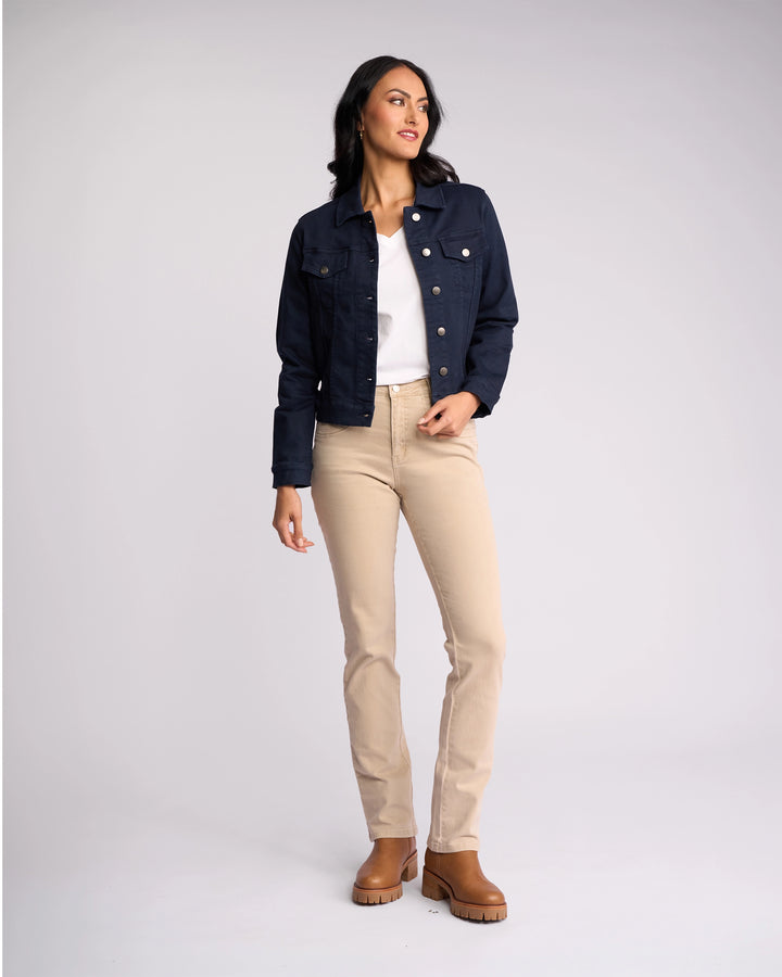 A woman stands against a plain background wearing the Vassalli Rider Knit Jacket - Carbon over a white shirt, beige pants, and brown ankle boots. She looks to the side with one hand in her pocket, showcasing the jacket’s stretch comfort.