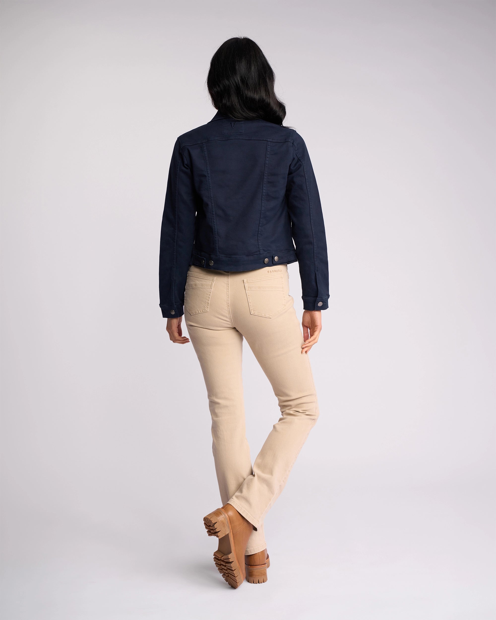 A woman faces away from the camera wearing the Vassalli Rider Knit Jacket in Carbon, paired with light beige pants and brown shoes, against a plain light background.