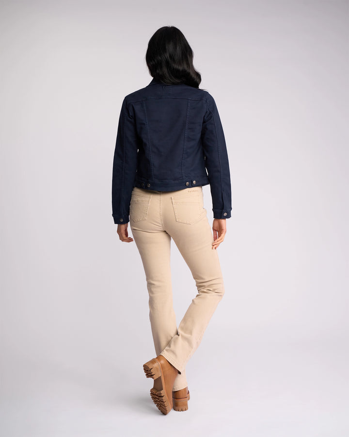 A woman faces away from the camera wearing the Vassalli Rider Knit Jacket in Carbon, paired with light beige pants and brown shoes, against a plain light background.