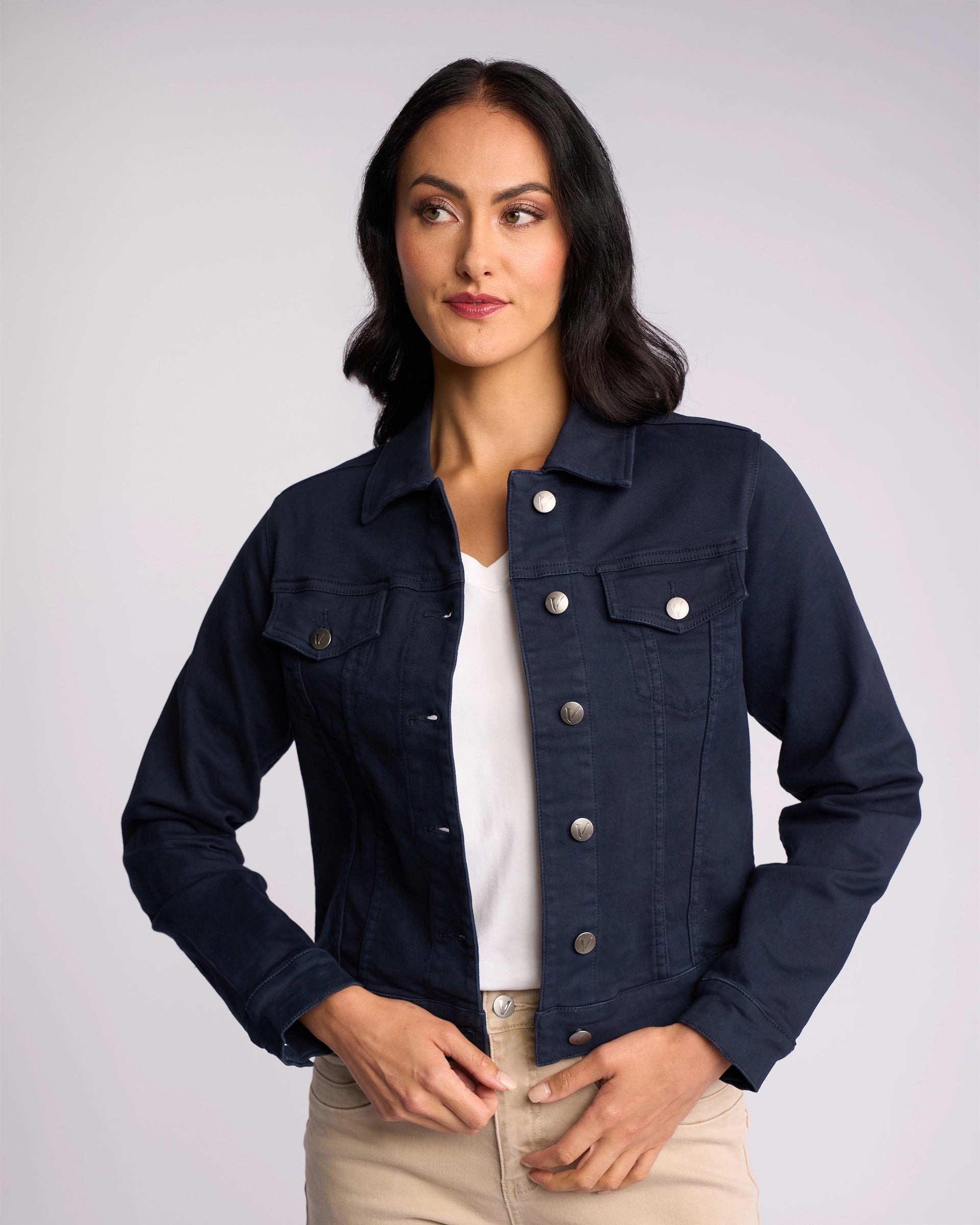 A woman with long dark hair wears a Vassalli Rider Knit Jacket - Carbon over a white shirt and beige pants. She stands against a plain light background, looking slightly to the side with a neutral expression.