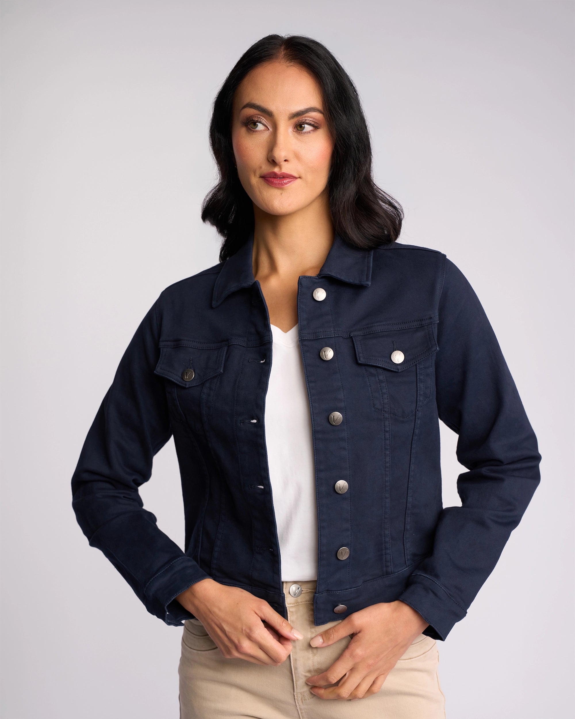 A woman with long dark hair wears a Vassalli Rider Knit Jacket - Carbon over a white shirt and beige pants. She stands against a plain light background, looking slightly to the side with a neutral expression.