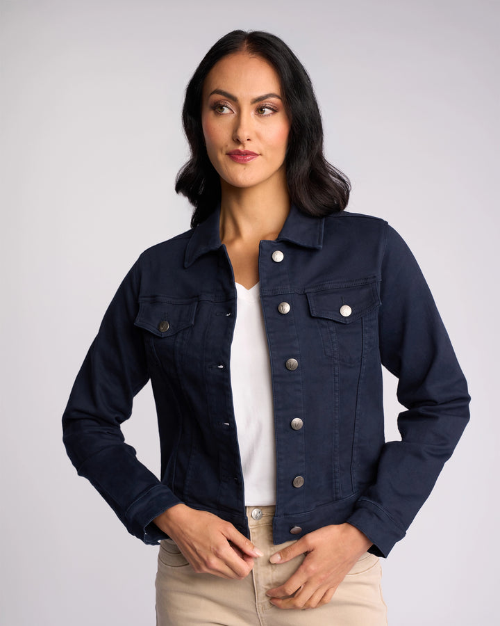 A woman with long dark hair wears a Vassalli Rider Knit Jacket - Carbon over a white shirt and beige pants. She stands against a plain light background, looking slightly to the side with a neutral expression.