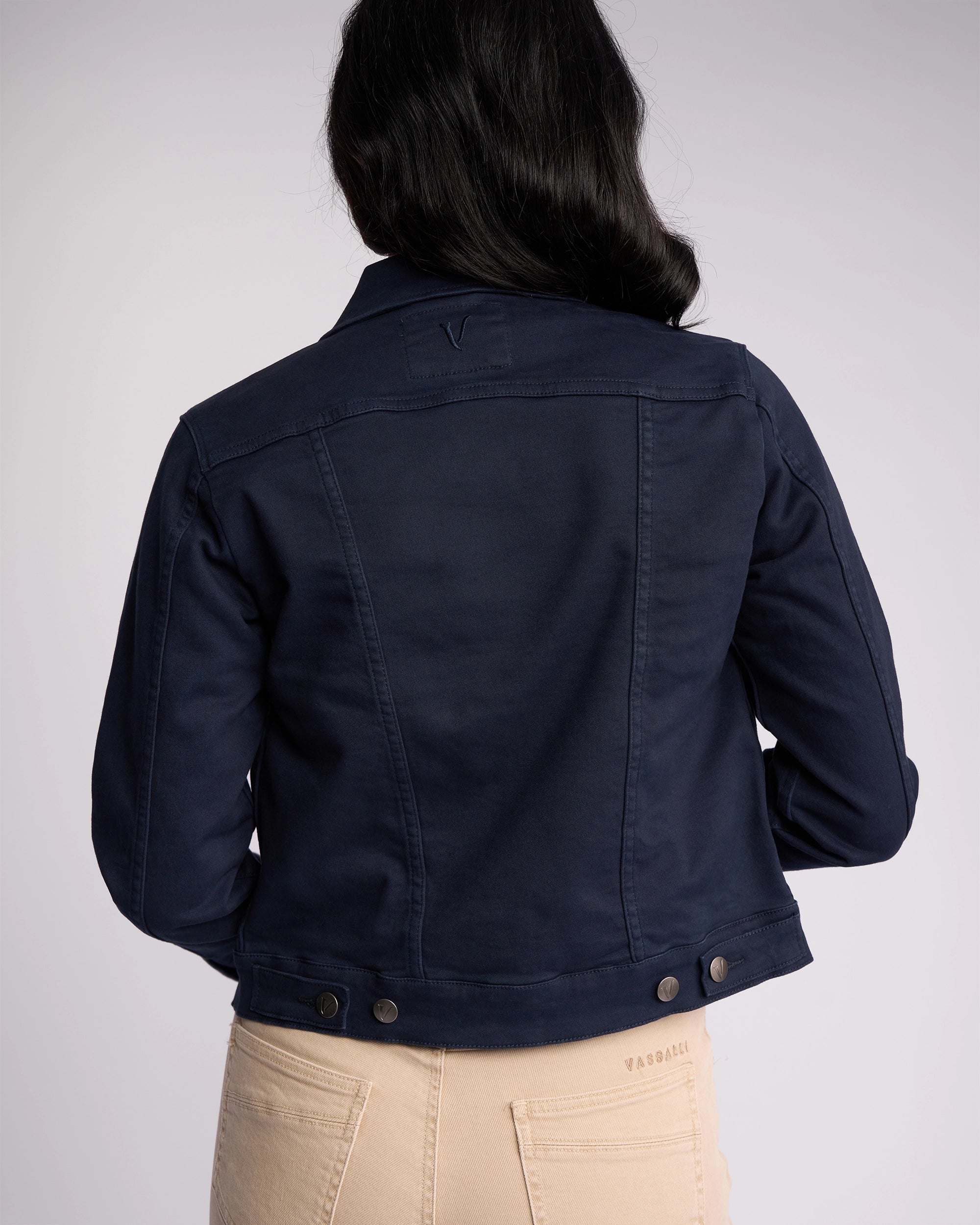 A person with long dark hair is shown from the back, wearing the Vassalli Rider Knit Jacket in Carbon and beige pants against a plain light background.