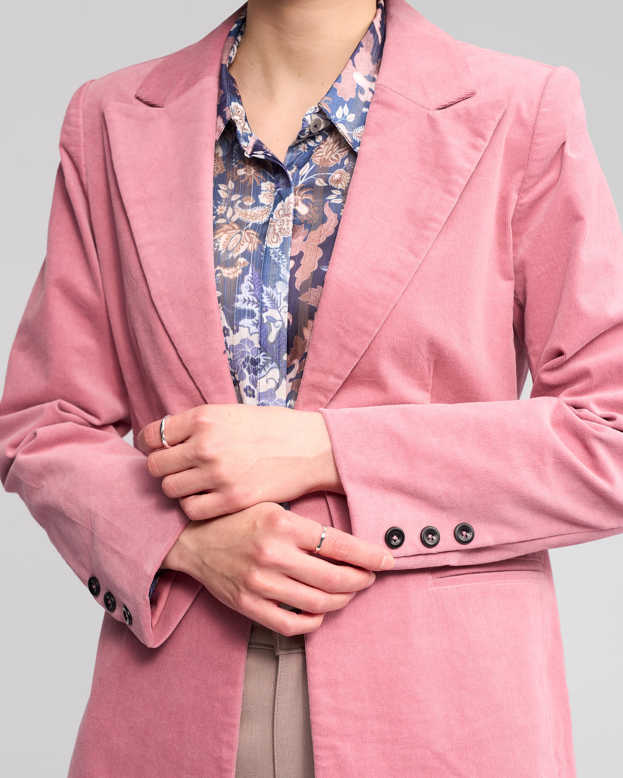 Wearing the Vassalli Gallery Blazer in dusky pink over a blue floral shirt, this person stands with hands clasped at the waist. Four buttons are visible on the blazer's sleeve; their face is not shown.