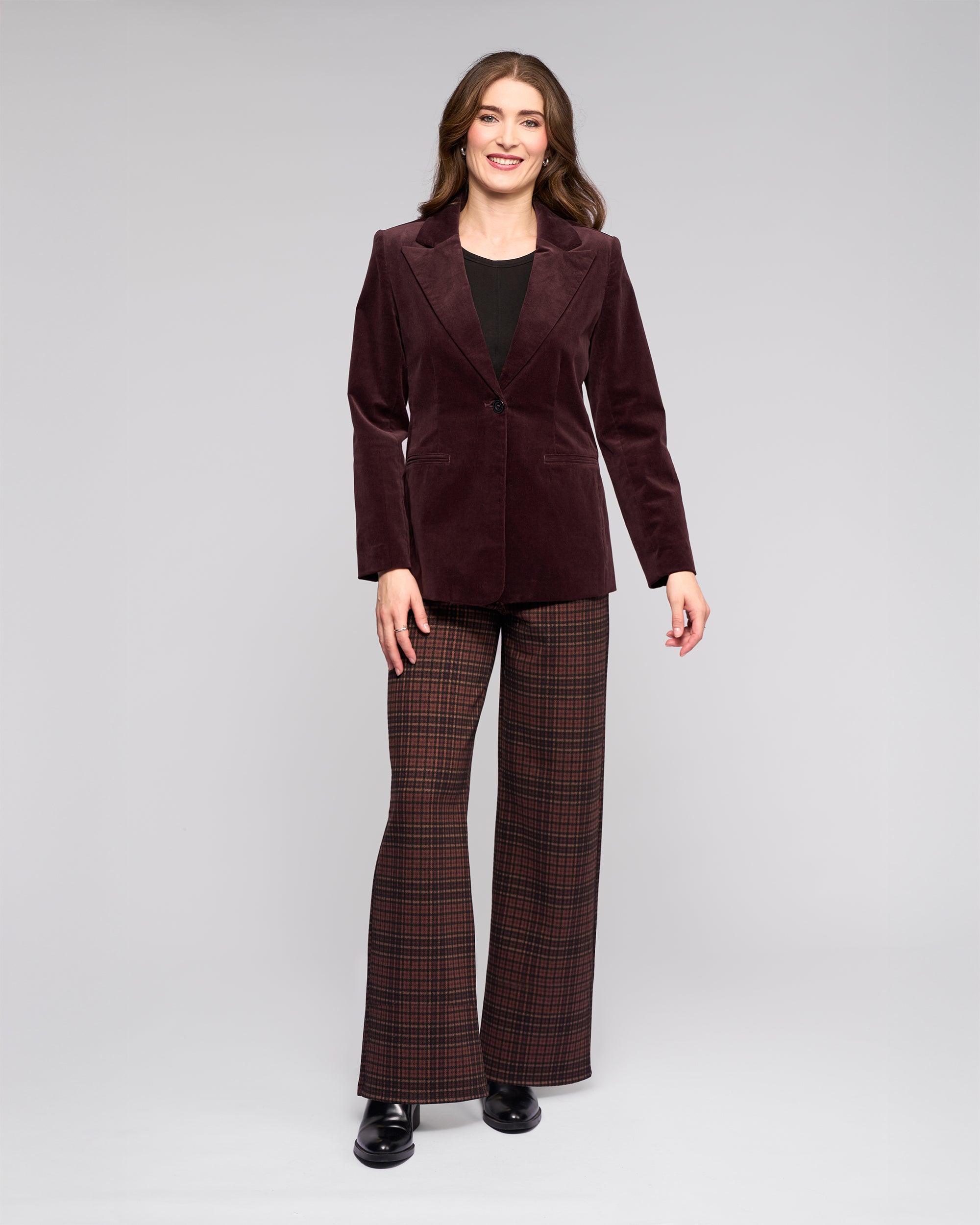 A woman smiles against a gray background, wearing the Vassalli Gallery Blazer in Merlot, a black top, brown and red plaid flared pants, and black shoes.