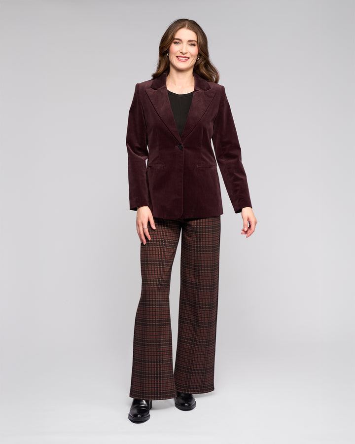 A woman smiles against a gray background, wearing the Vassalli Gallery Blazer in Merlot, a black top, brown and red plaid flared pants, and black shoes.