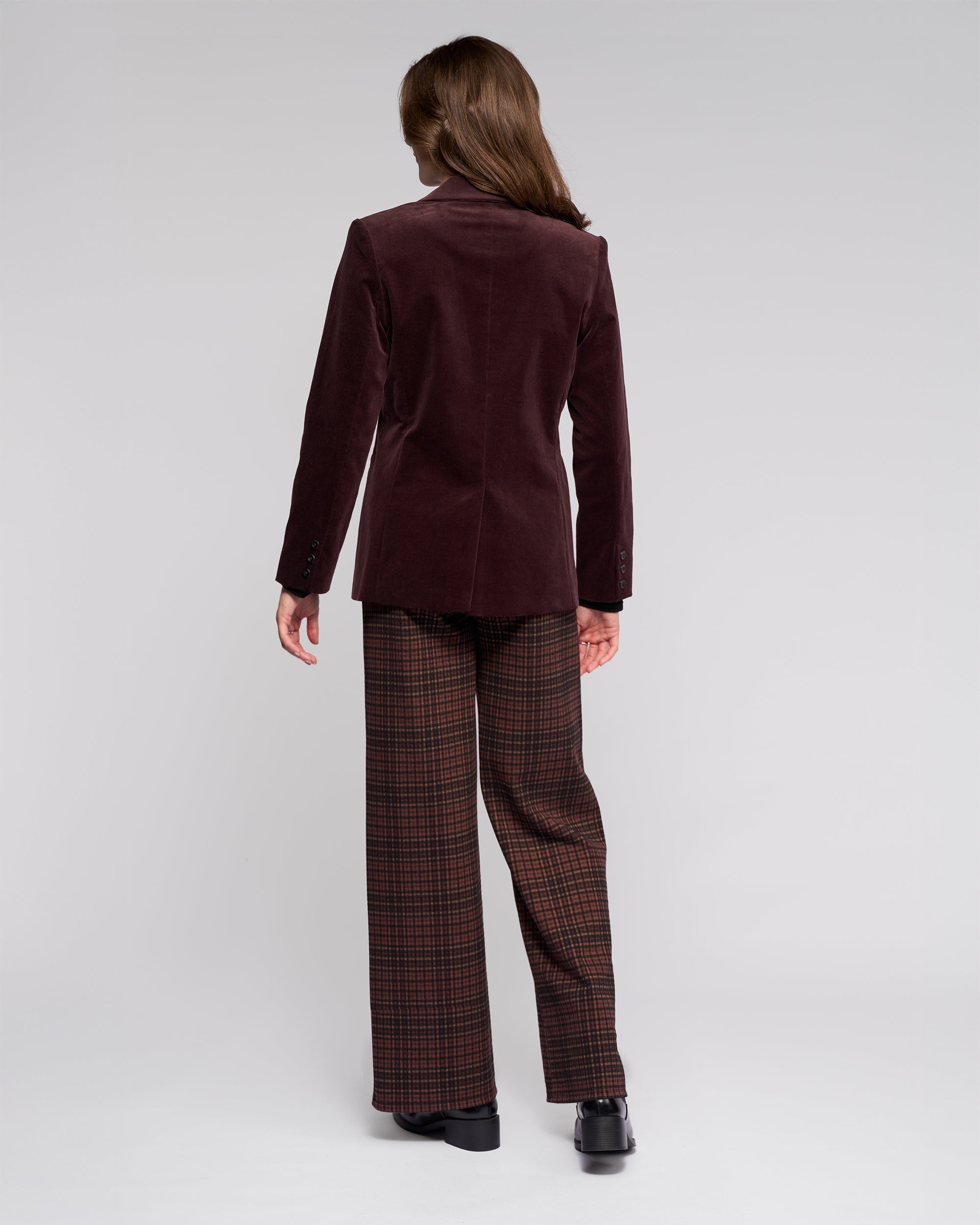 A person with long brown hair stands with their back to the camera, wearing the Vassalli Gallery Blazer in Merlot, plaid brown wide-leg pants, and black shoes against a plain light gray background.