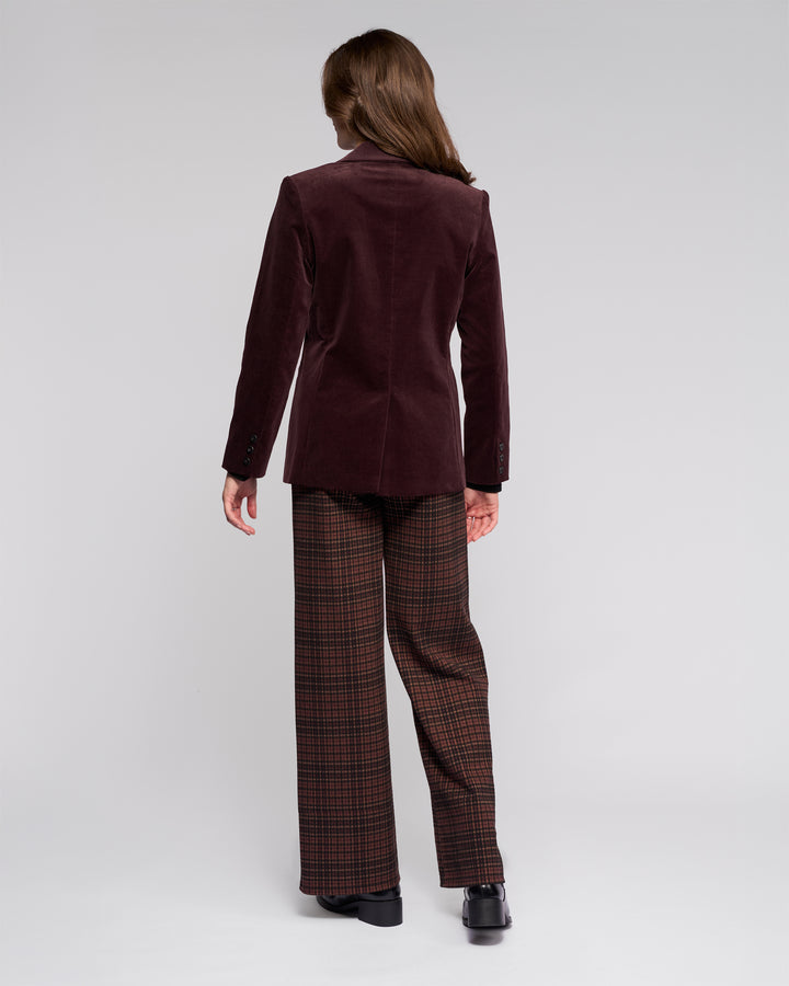 A person with long brown hair stands with their back to the camera, wearing the Vassalli Gallery Blazer in Merlot, plaid brown wide-leg pants, and black shoes against a plain light gray background.