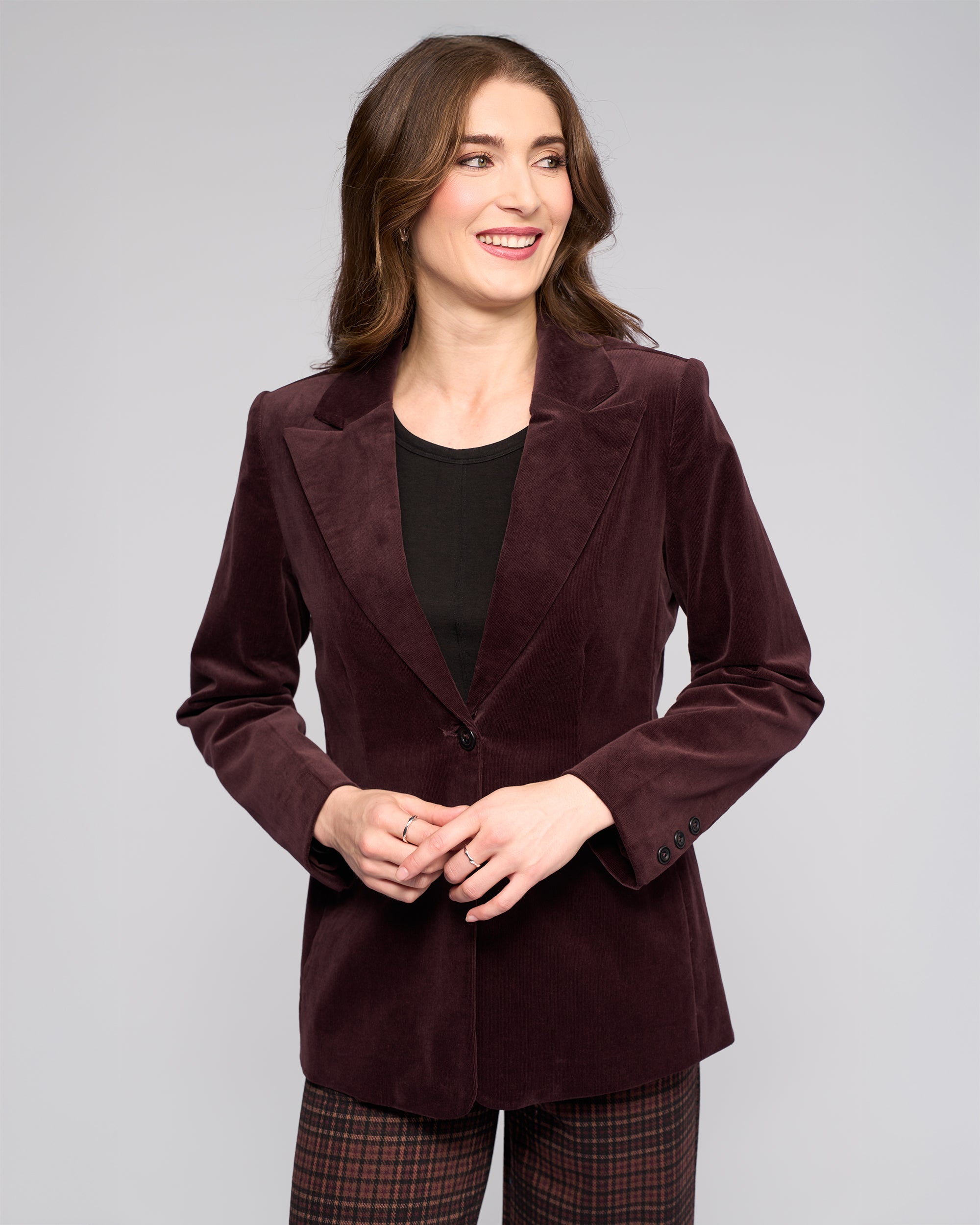 A woman with long brown hair wears the Vassalli Gallery Blazer in Merlot over a black top and plaid pants, standing against a light gray background, smiling and looking to her left.