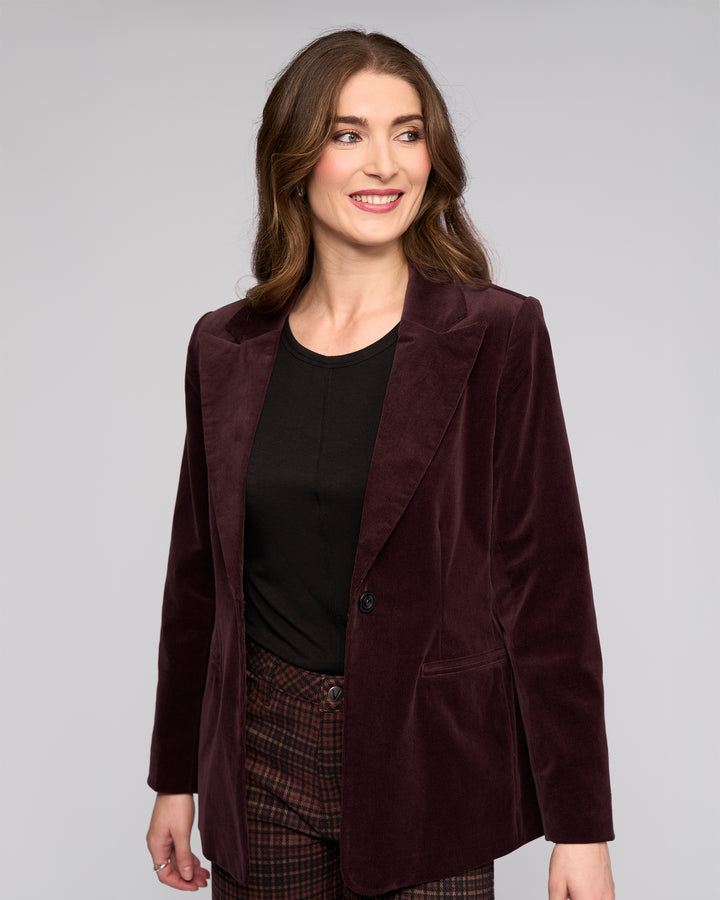 A woman with long brown hair smiles and looks left, wearing the Vassalli Gallery Blazer in Merlot over a black top and plaid pants against a plain light gray background.