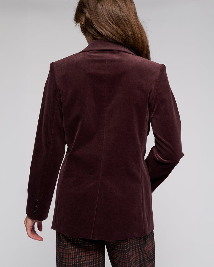 A person with long brown hair is shown from behind, wearing the Vassalli Gallery Blazer in Merlot and plaid pants against a plain light background.