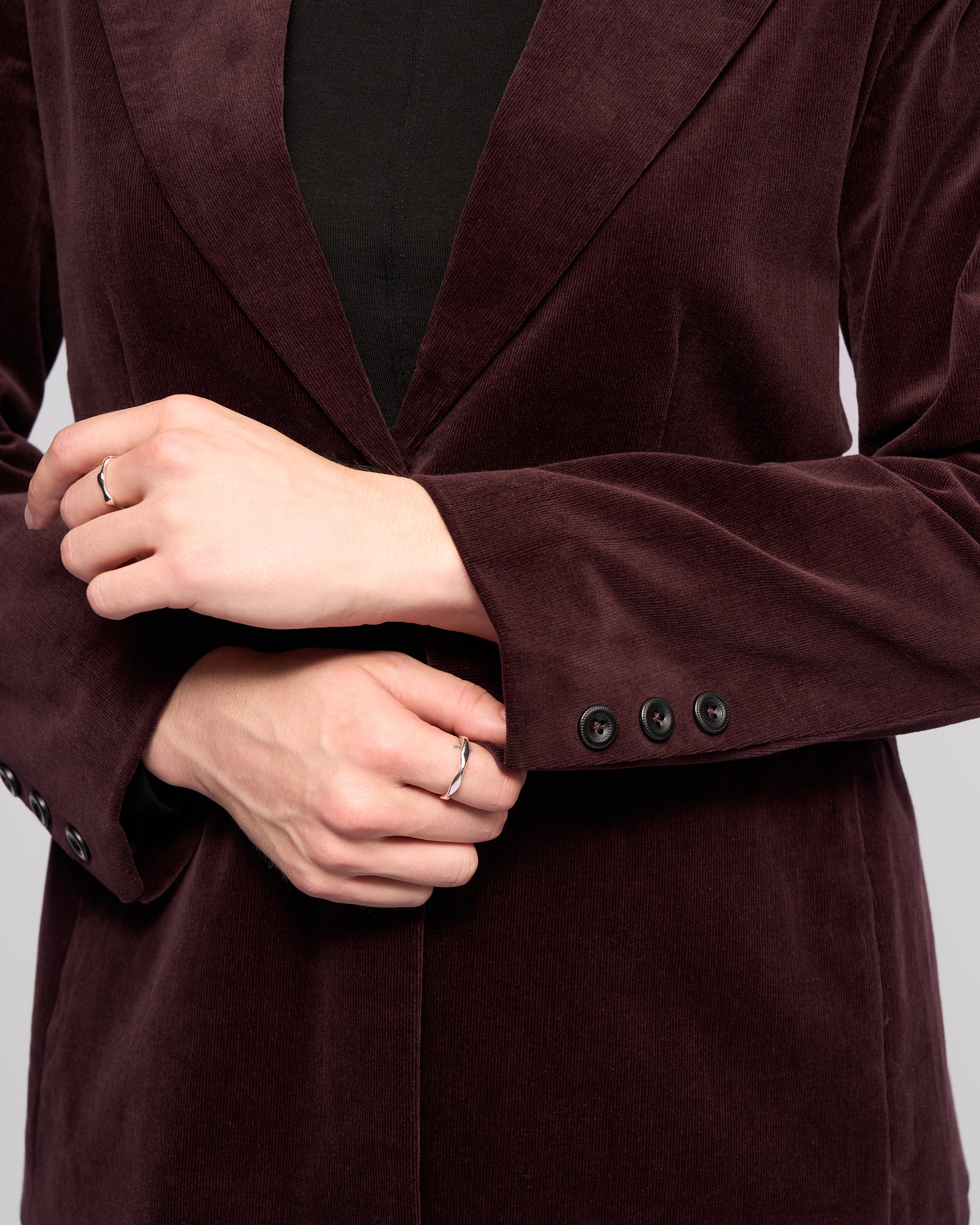 A person stands with hands clasped, displaying silver rings and black sleeve buttons while wearing the Vassalli Gallery Blazer in Merlot over a black top.