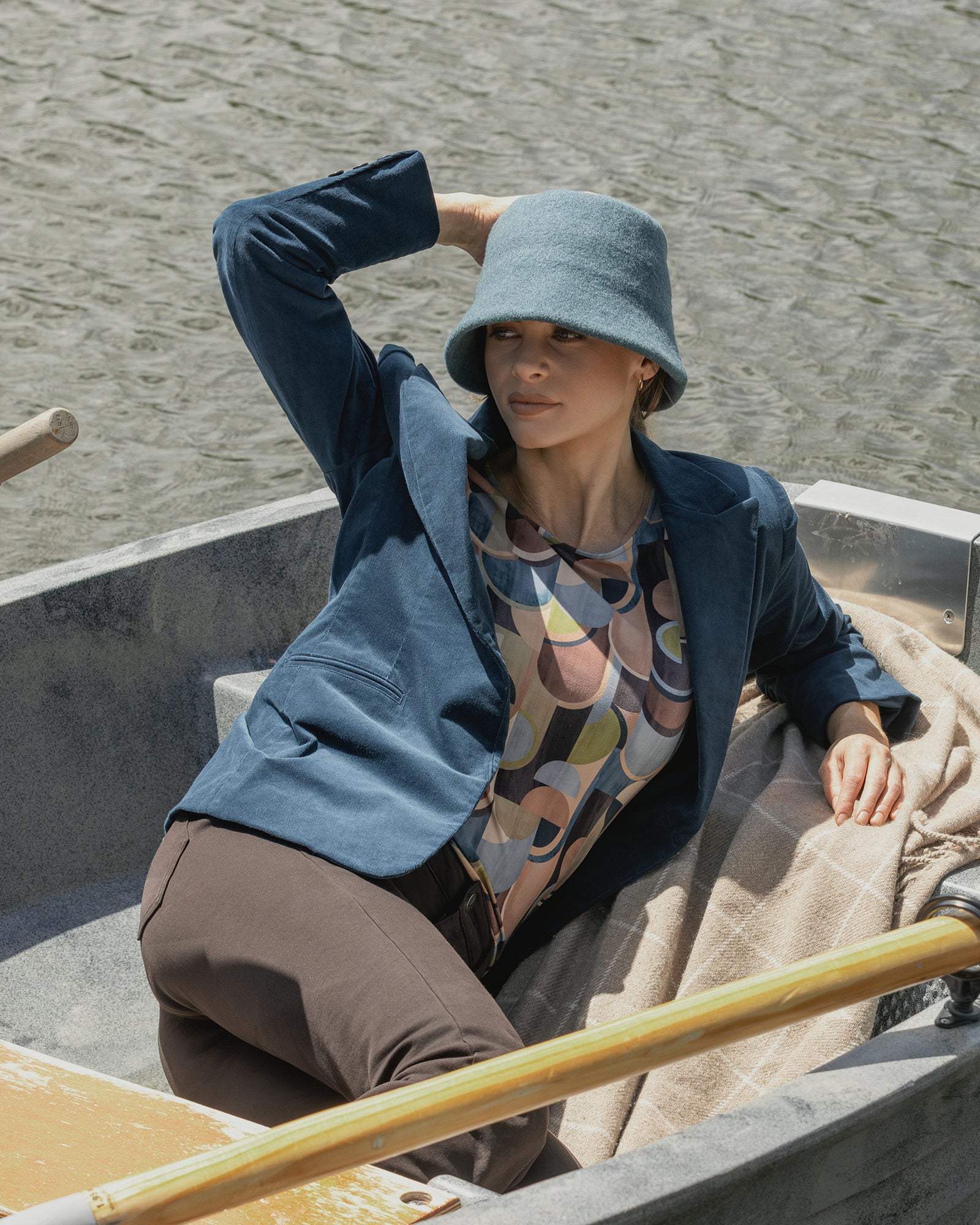 A woman in the Vassalli Gallery Blazer - Petrol and a dark teal cord bucket hat lounges in a rowboat, one arm on her head, the other on the boat’s edge, with a patterned shirt and blanket by her side.