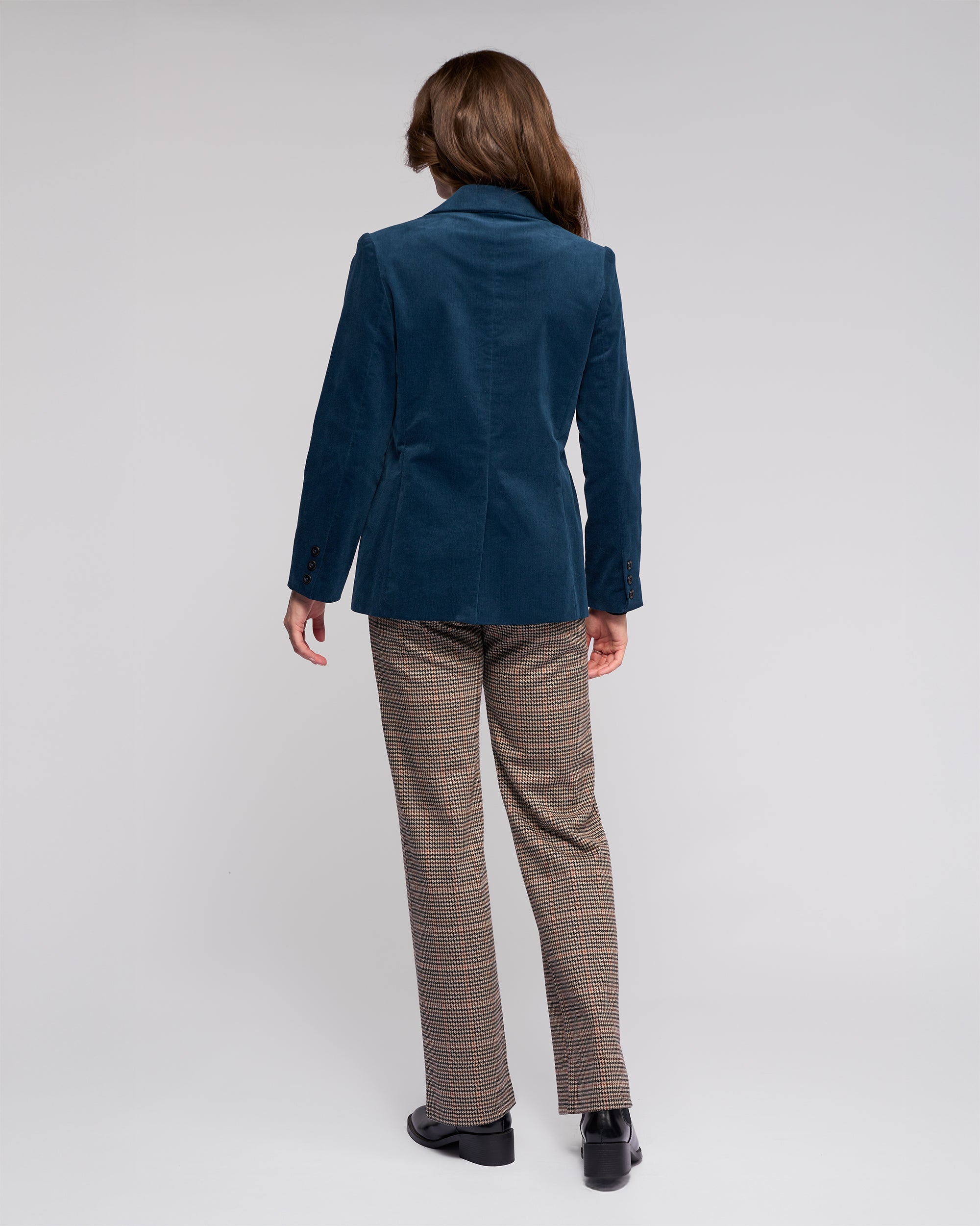 A person with long brown hair, seen from behind, wears the Vassalli Gallery Blazer in Petrol, pinwale cord plaid brown trousers, and black shoes, standing against a plain light gray background.