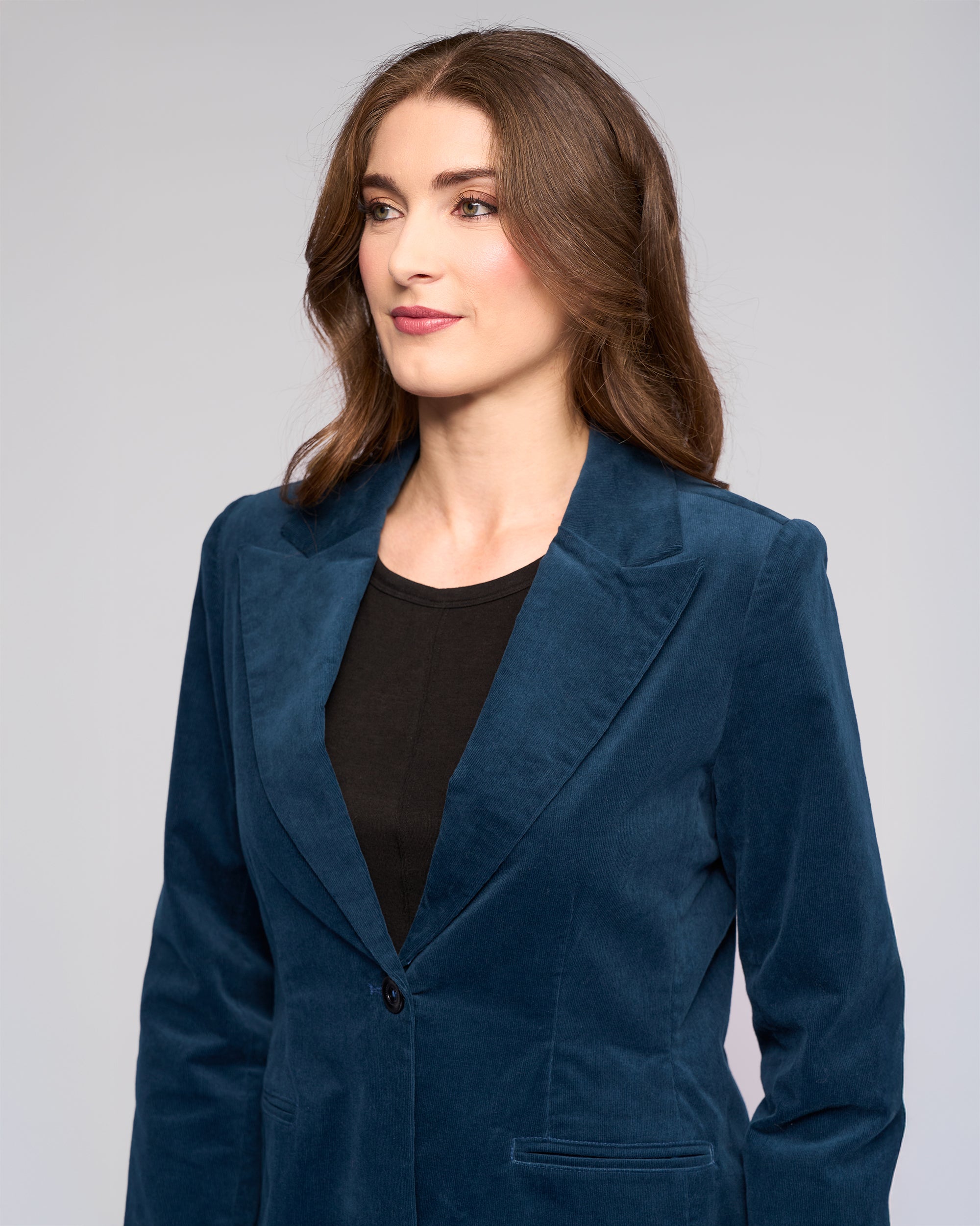A woman with long brown hair models the Vassalli Gallery Blazer in Petrol over a black top, glancing to the side against a plain light gray background.