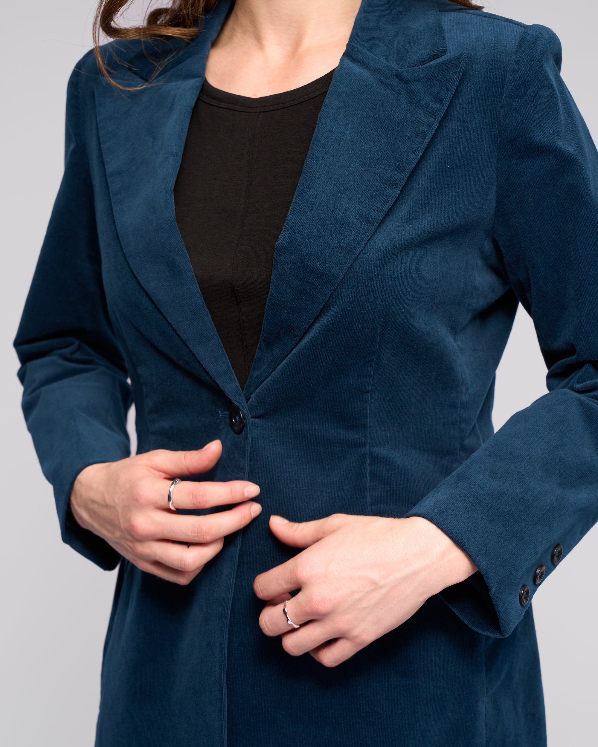 A person adjusts the button on the Vassalli Gallery Blazer in Petrol, worn over a black top. Only their upper body is visible, with their face partially out of frame.