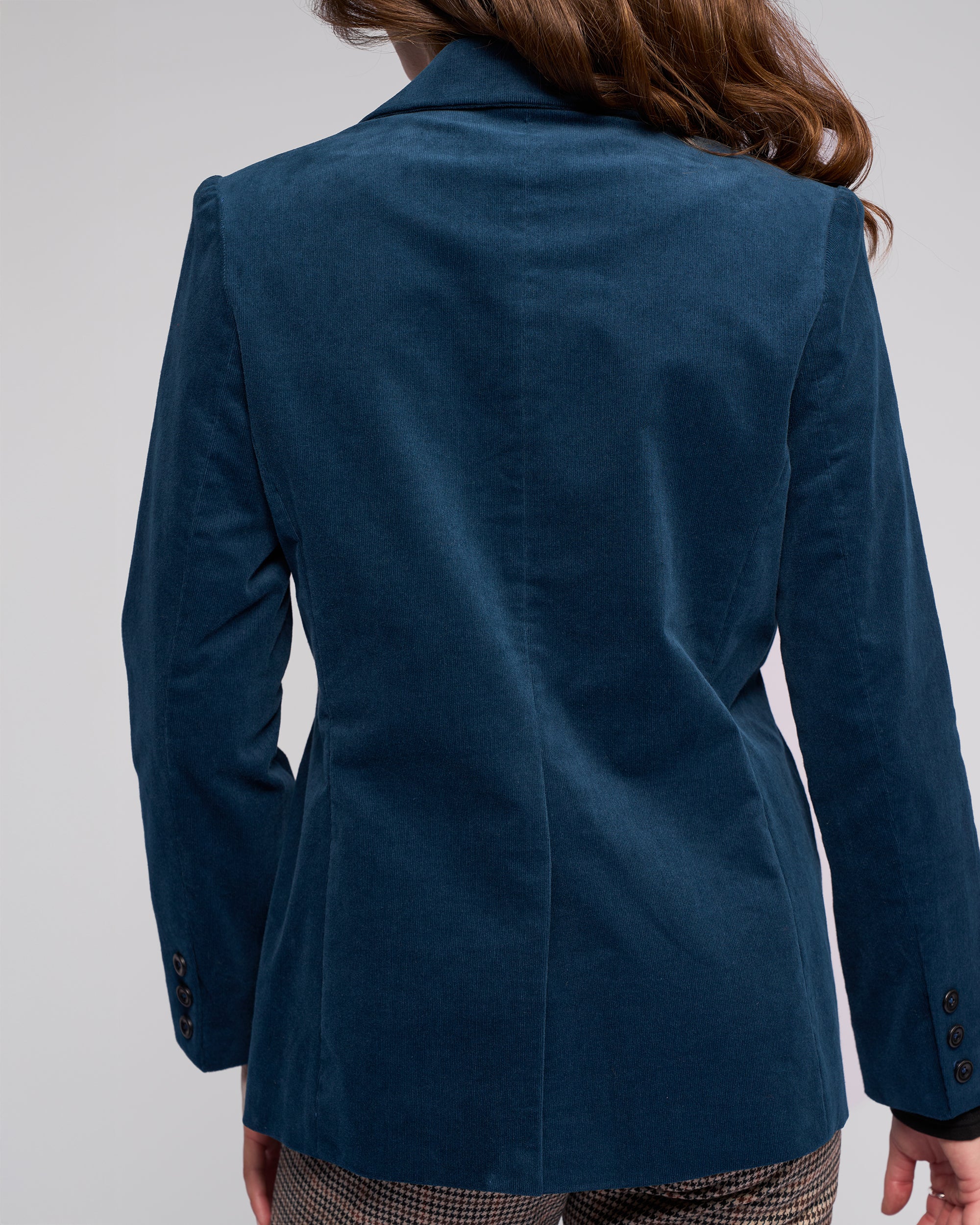 A person with long brown hair is seen from behind wearing the Vassalli Gallery Blazer in Petrol and brown plaid pants against a plain light background.