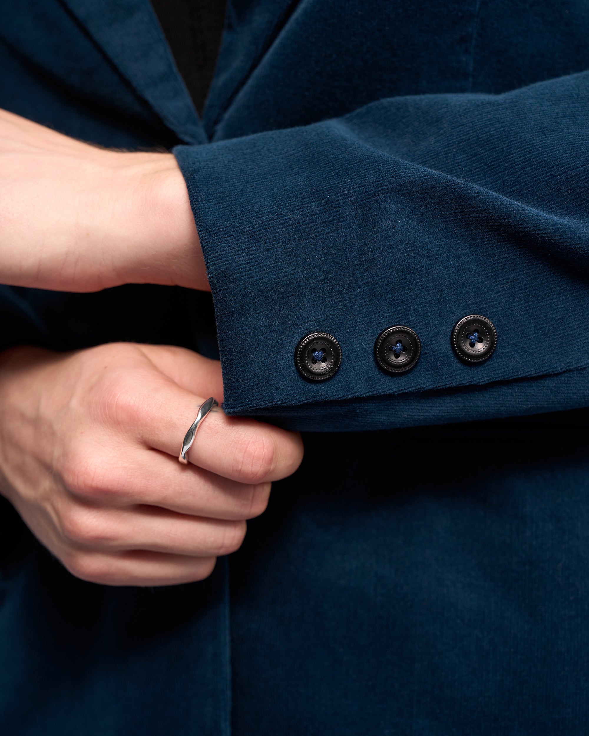 A close-up of a hand adjusting the sleeve of the Vassalli Gallery Blazer - Petrol, a dark teal semi-tailored jacket with three black buttons; the hand wears a silver ring.