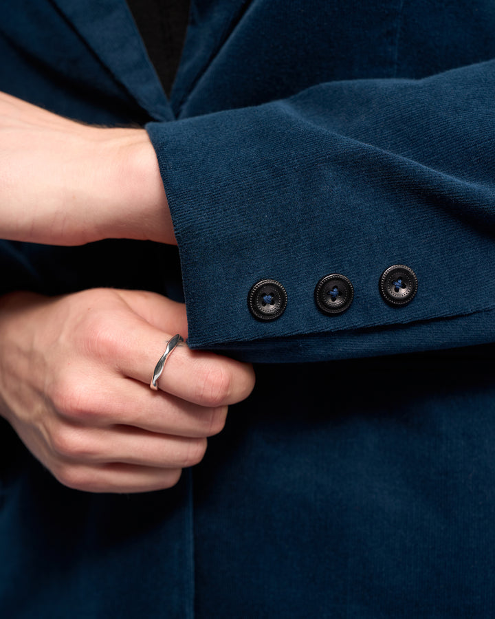 A close-up of a hand adjusting the sleeve of the Vassalli Gallery Blazer - Petrol, a dark teal semi-tailored jacket with three black buttons; the hand wears a silver ring.