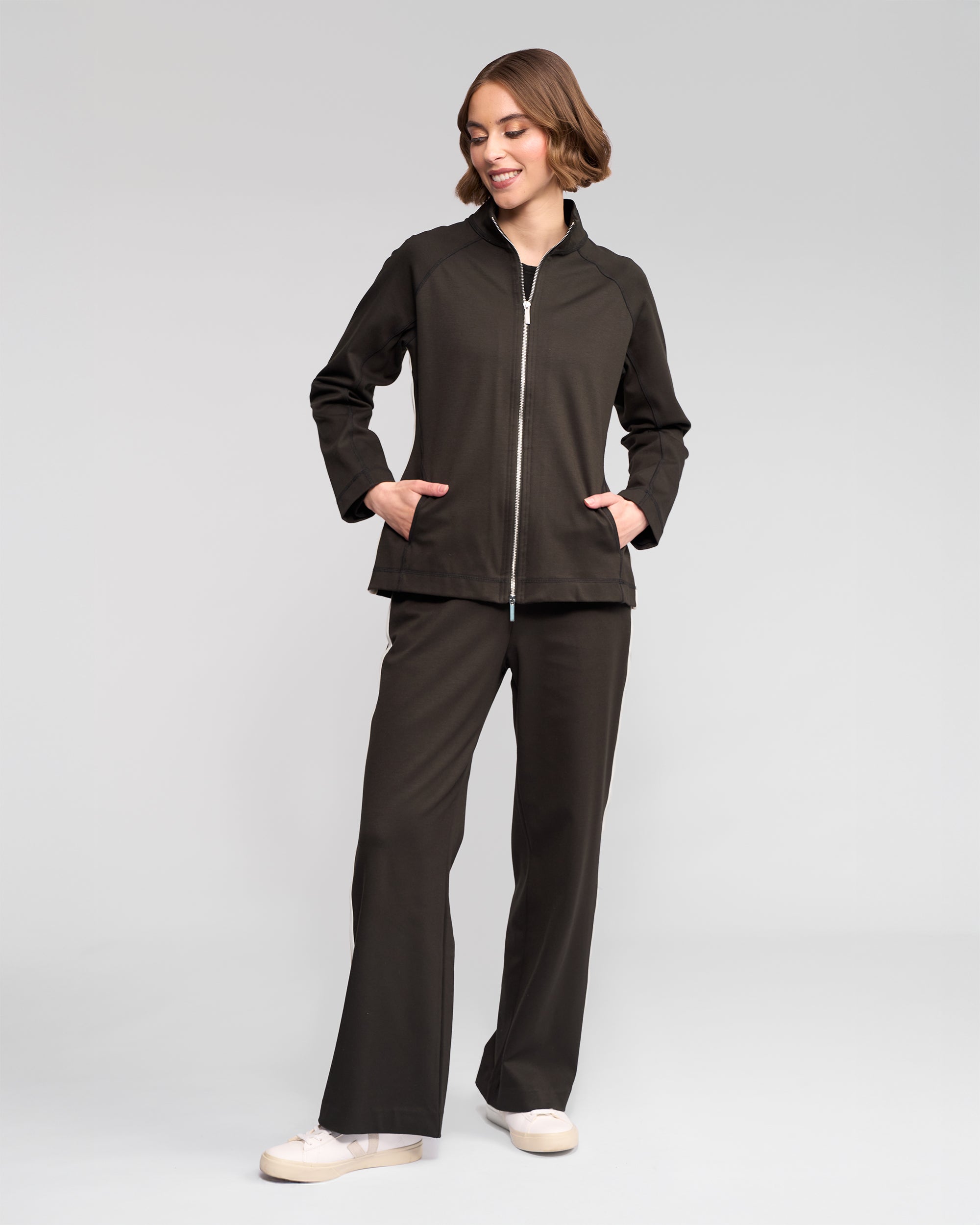A person smiles and looks to the side with hands in pockets, wearing the Vassalli Weekend Top - Dark Hunter and matching pants against a plain light gray background, paired with cream-colored sneakers.