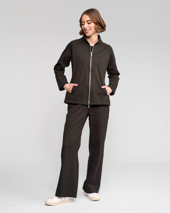 A person smiles and looks to the side with hands in pockets, wearing the Vassalli Weekend Top - Dark Hunter and matching pants against a plain light gray background, paired with cream-colored sneakers.