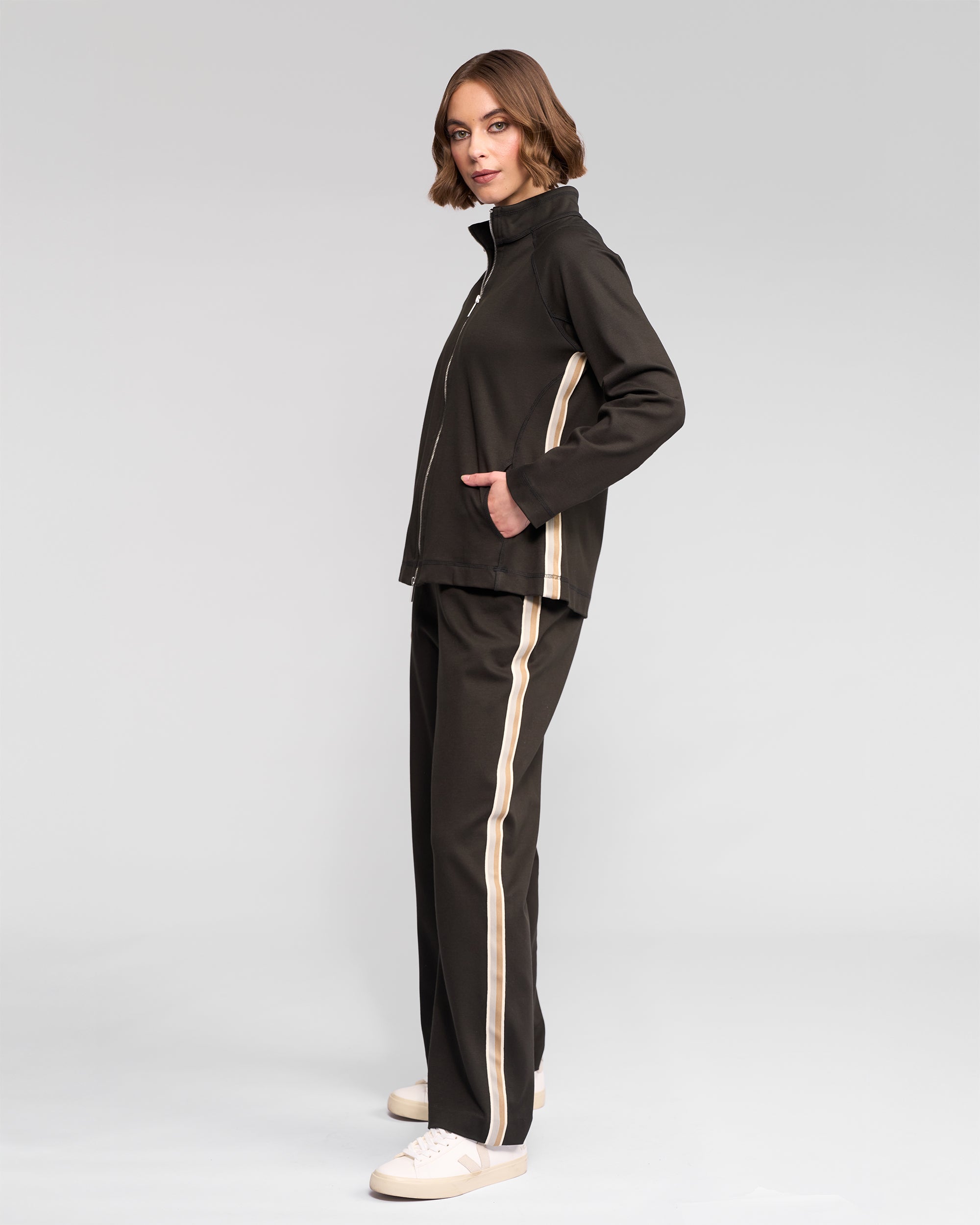 A woman with short, wavy brown hair models the Vassalli Weekend Top - Dark Hunter jacket with beige side stripes, hands in pockets and white sneakers, against a plain light background, looking at the camera.