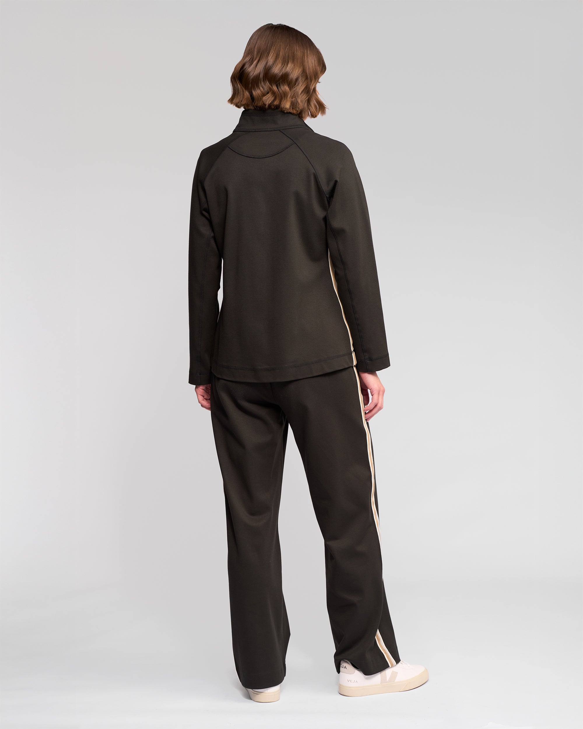 A person with short, wavy hair faces away from the camera, wearing the Vassalli Weekend Top in Dark Hunter with beige side stripes and white sneakers, standing against a plain light gray background.