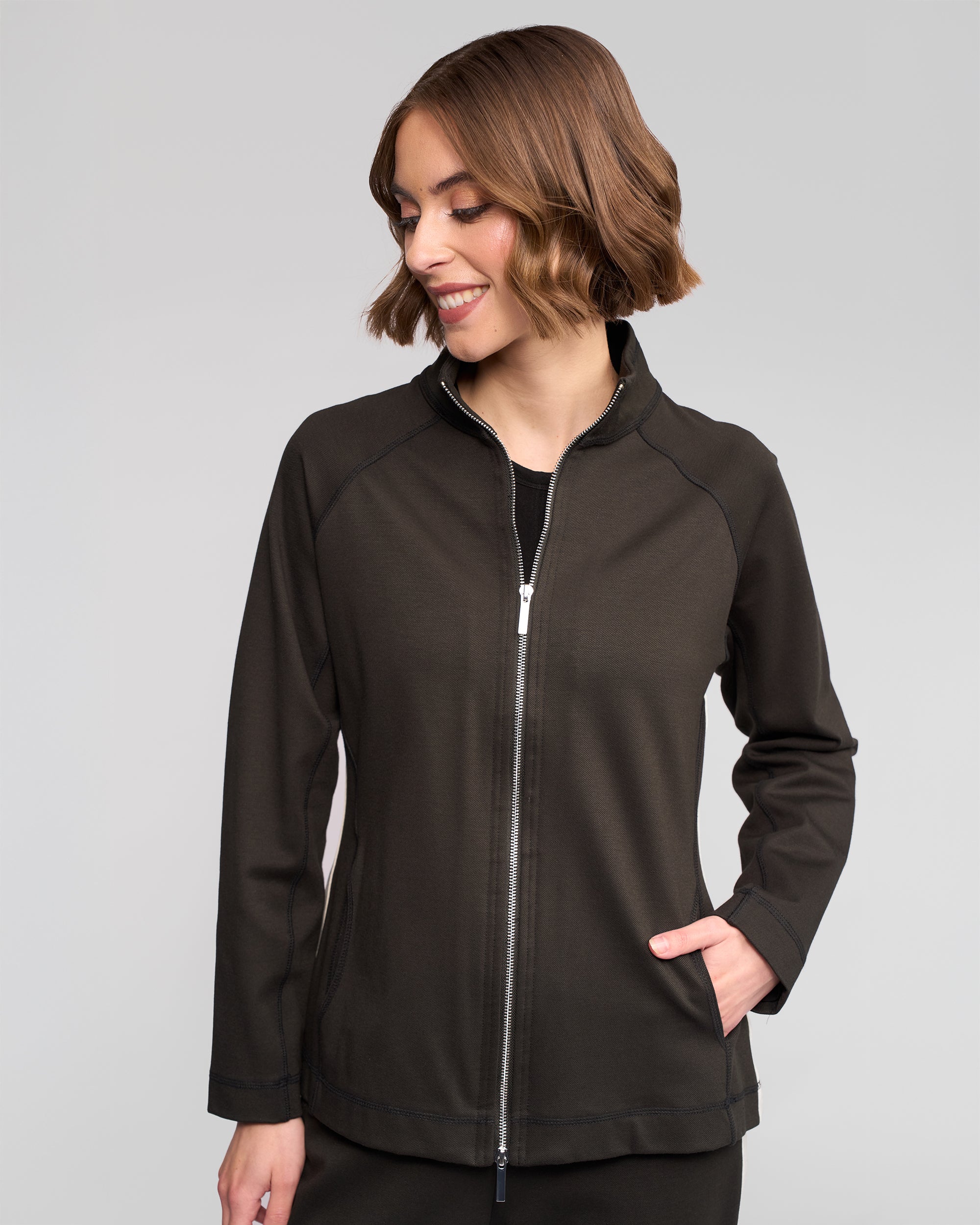 A woman with short brown hair smiles while looking down, wearing the Vassalli Weekend Top in Dark Hunter with her right hand in the pocket, standing against a plain light gray background.