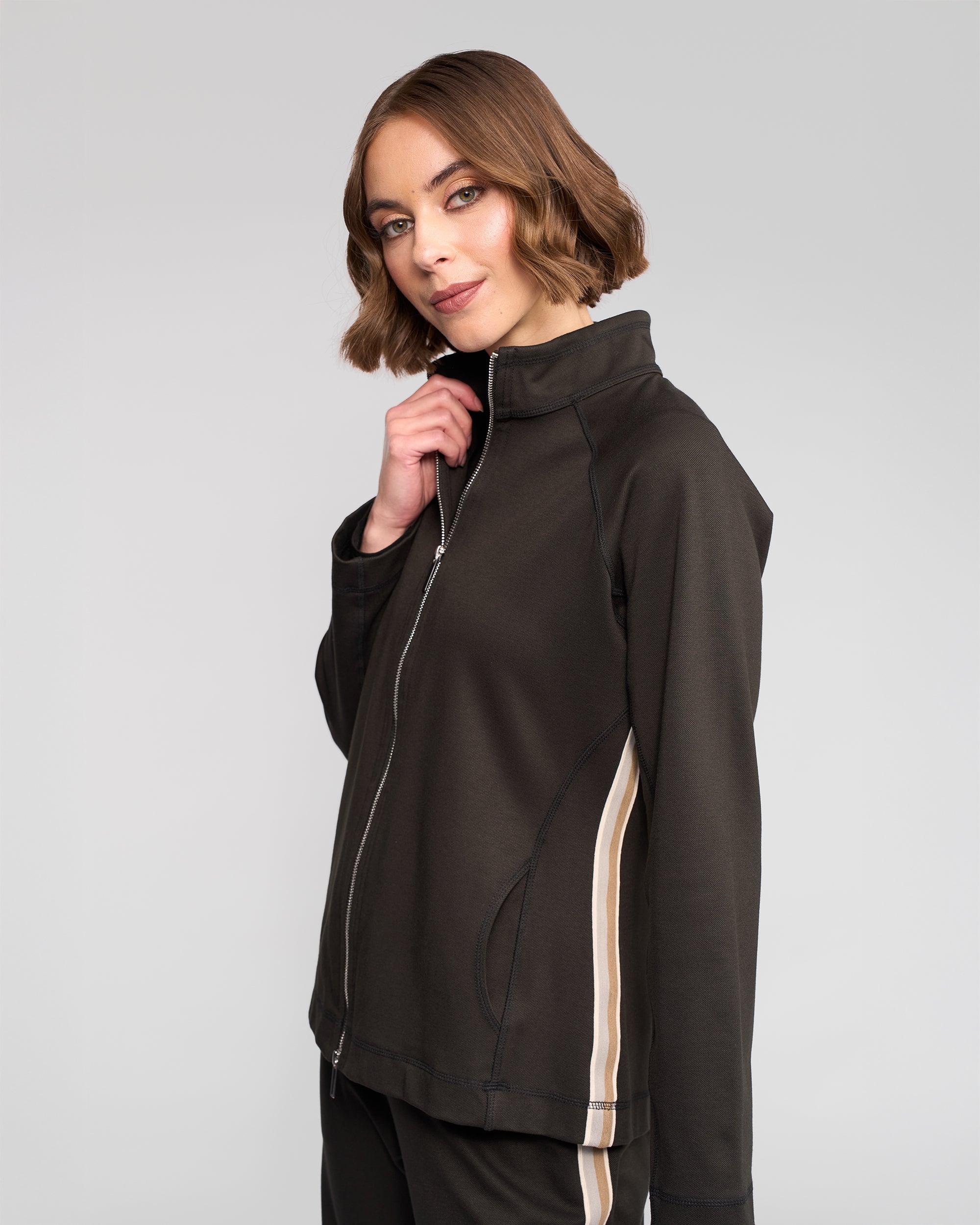 A woman with short brown hair confidently poses against a plain light gray background, wearing the Vassalli Weekend Top - Dark Hunter with beige side stripes.