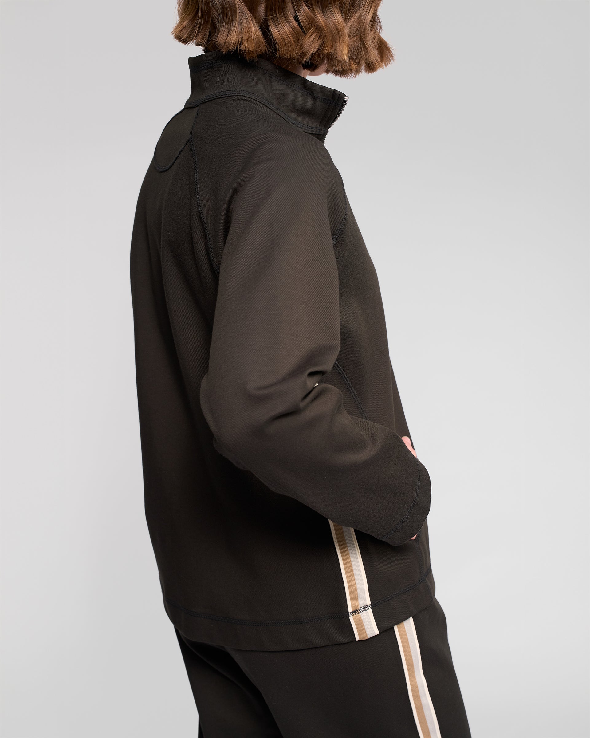 A person with short brown hair, wearing the Vassalli Weekend Top in Dark Hunter, stands with one hand in their pocket and faces away from the camera.