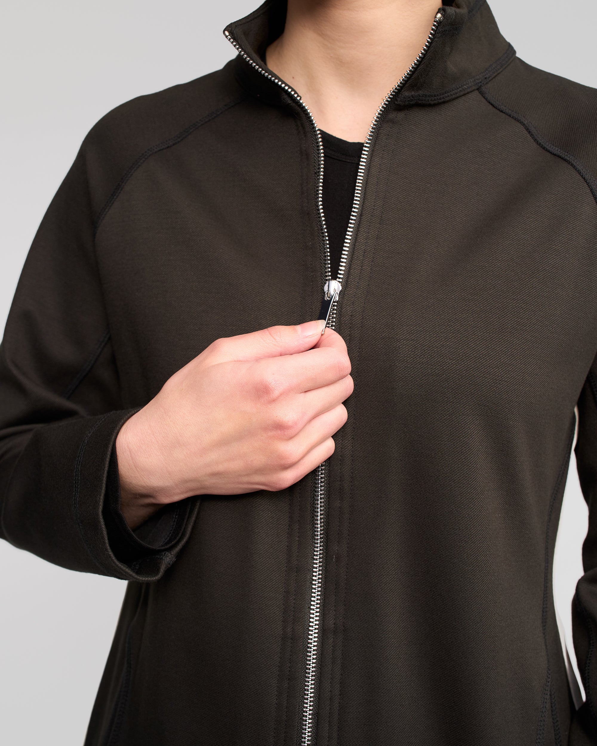 A person is shown from shoulders to waist, zipping up the Vassalli Weekend Top - Dark Hunter against a plain, light background.