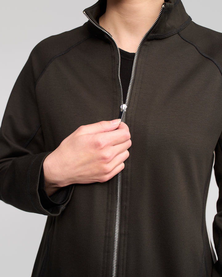 A person is shown from shoulders to waist, zipping up the Vassalli Weekend Top - Dark Hunter against a plain, light background.