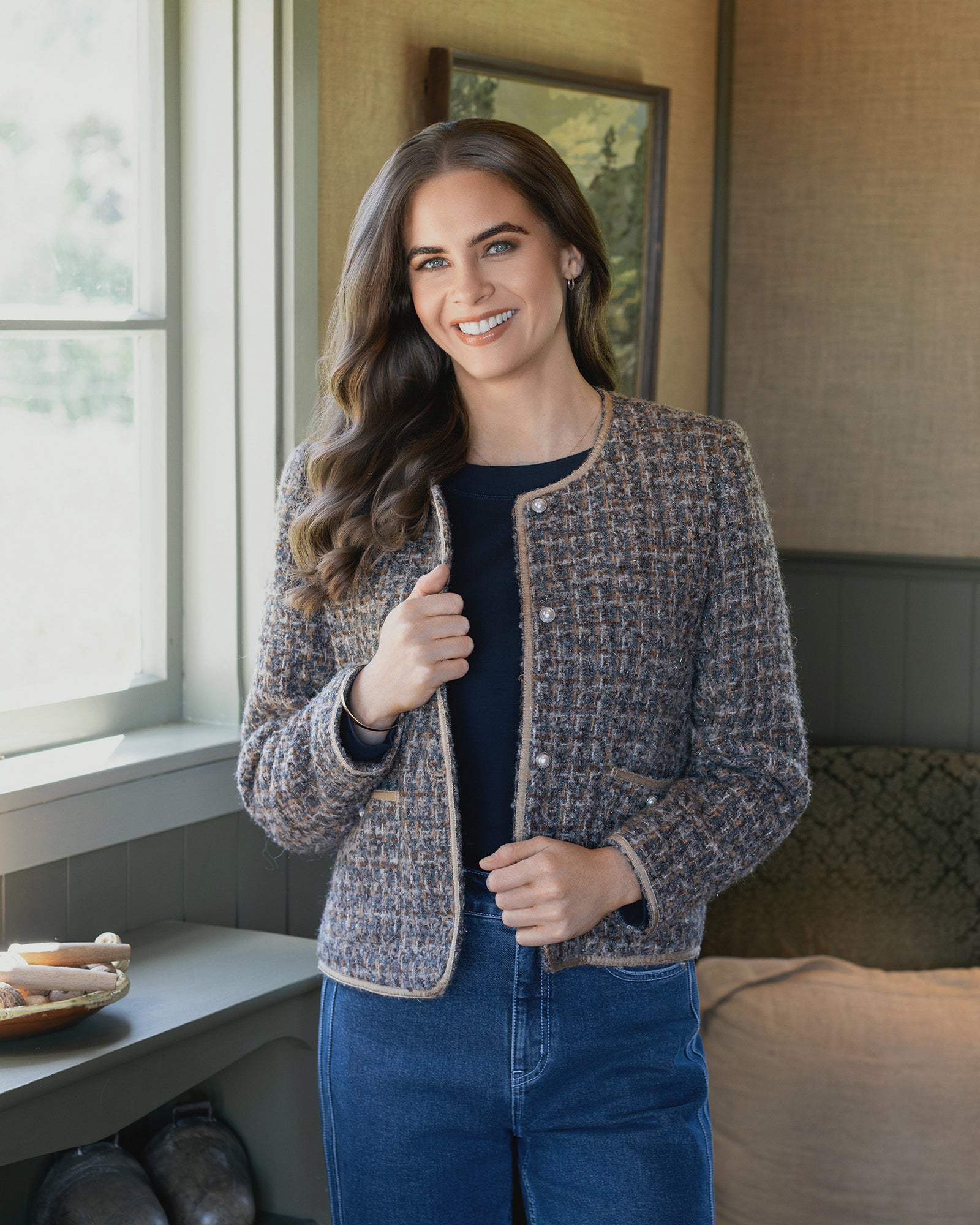 A woman with long brown hair smiles at the camera indoors, wearing the Vassalli Stage Jacket - Florence and blue jeans. A framed picture hangs on the wall, and a couch is visible in the background.