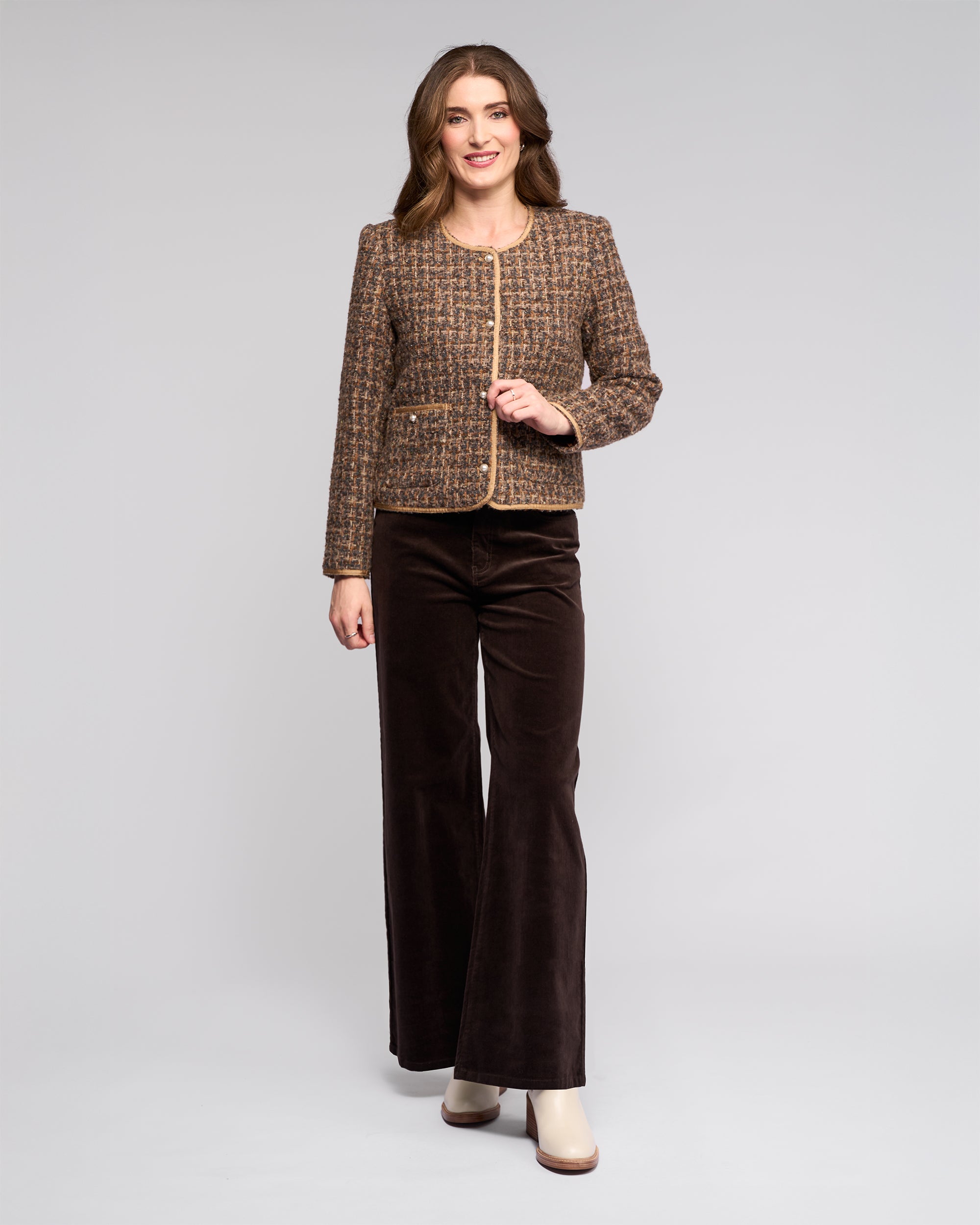 A woman with long, wavy brown hair smiles against a light gray background, wearing the Vassalli Stage Jacket - Florence, paired with dark brown wide-leg pants and cream-colored shoes.