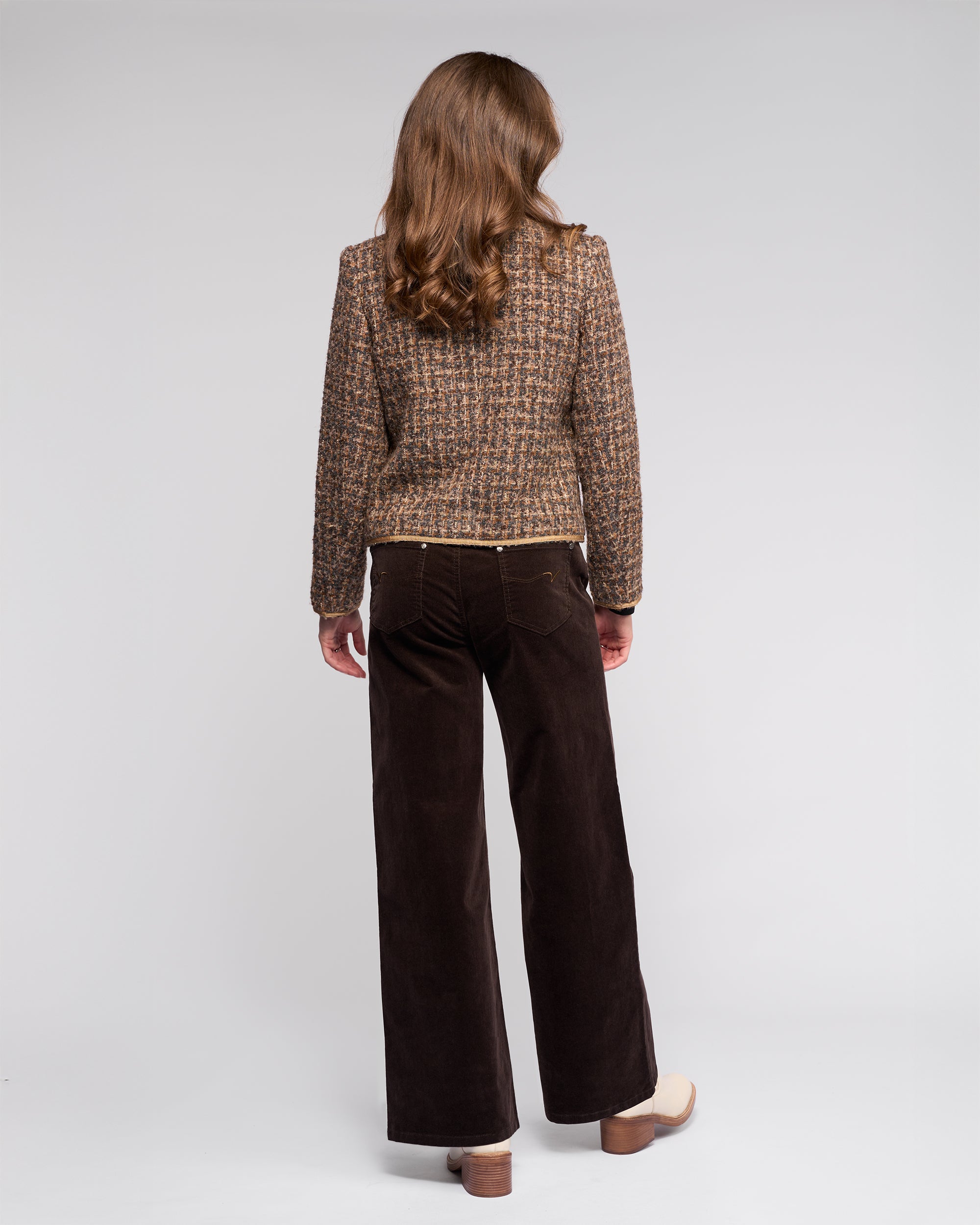A woman with long brown hair, seen from behind, wears the Vassalli Stage Jacket - Florence with dark brown wide-leg pants and beige platform shoes against a plain light background.
