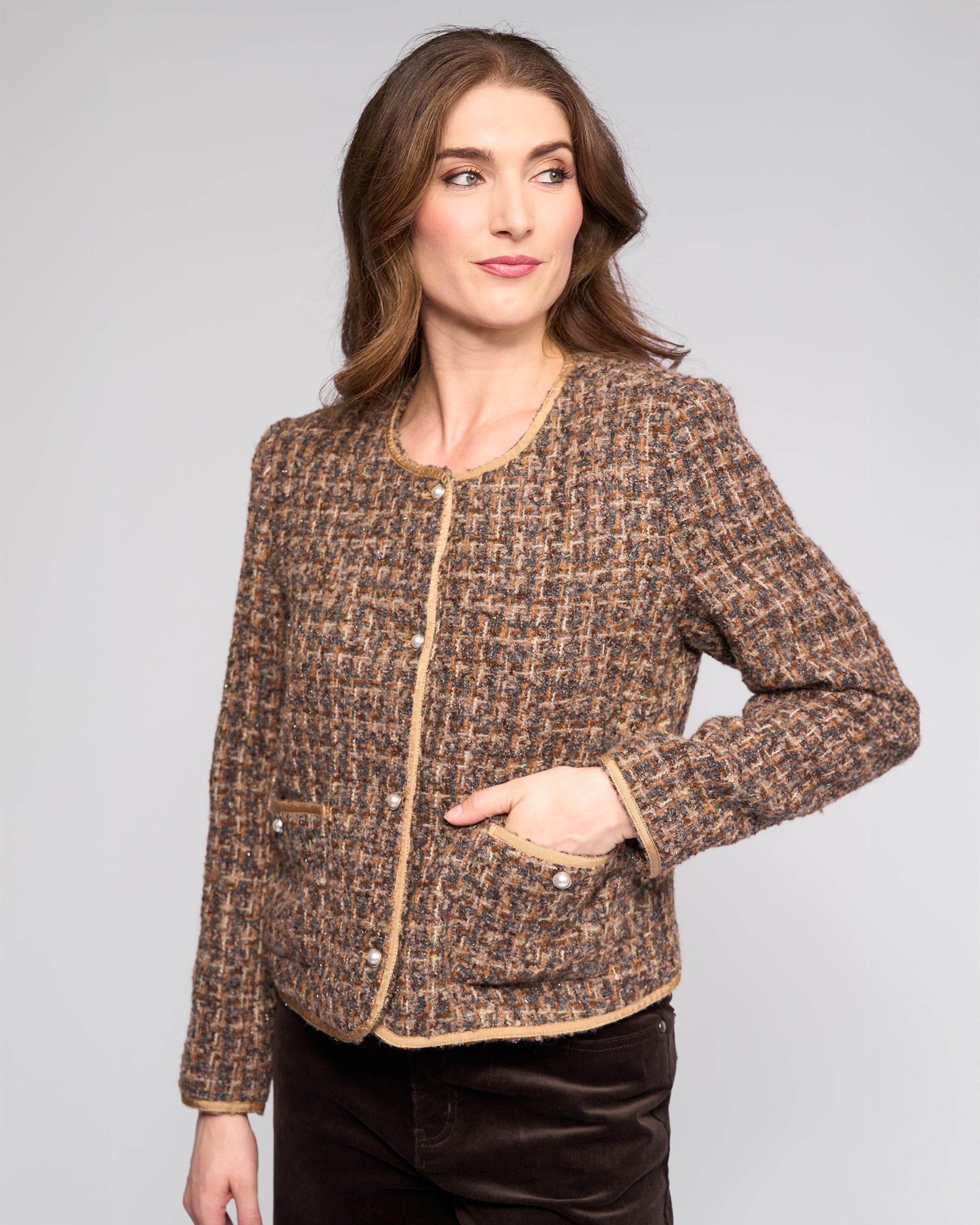 A woman with wavy brown hair wears the Vassalli Stage Jacket - Florence, a collarless textured woven style with beige trim and button details, paired with dark pants, standing against a plain light gray background.