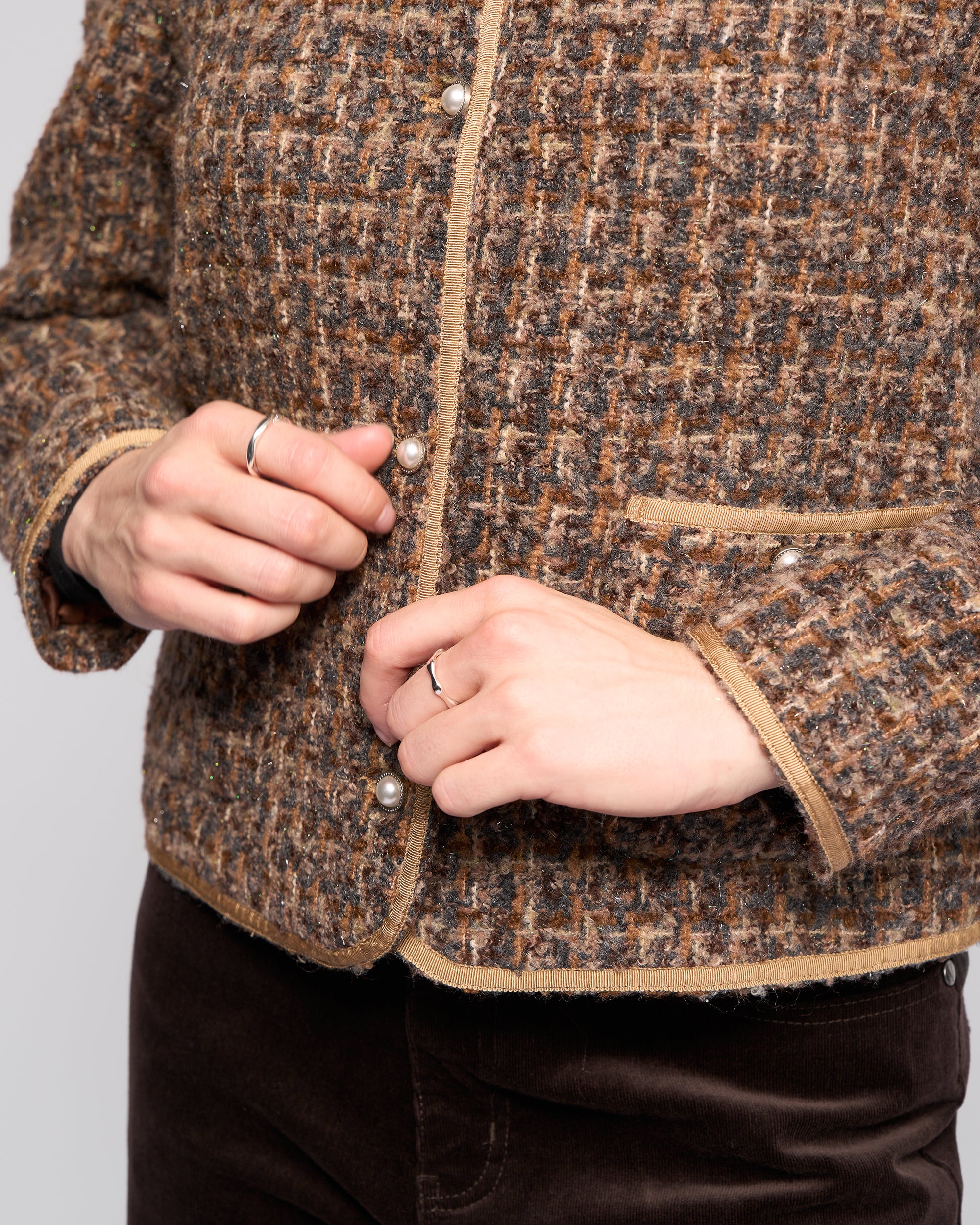 A person is fastening a button on the Vassalli Stage Jacket - Florence, a collarless textured woven design with beige trim and pearl buttons. Only their torso and ring-adorned hands are visible.