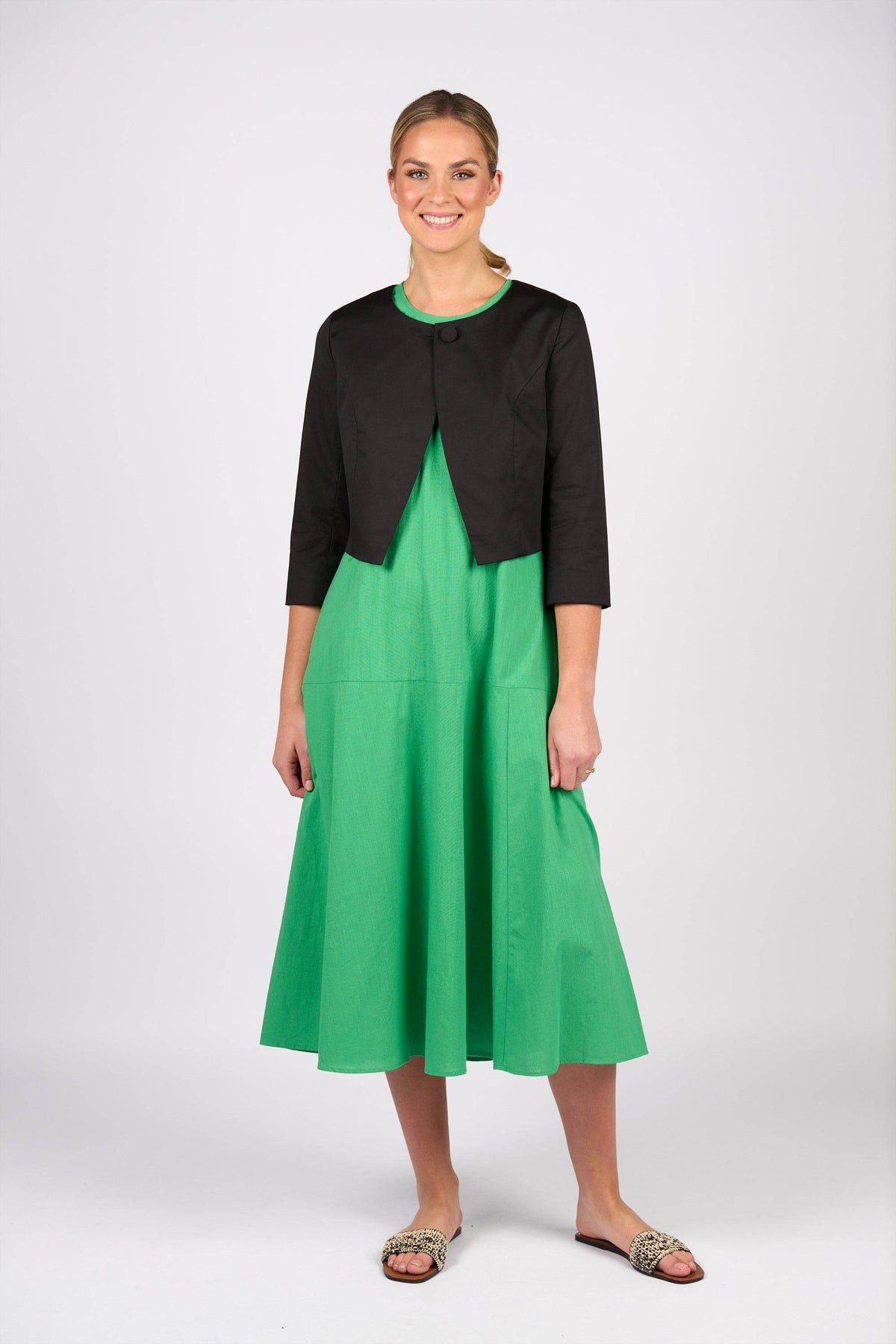 A woman stands smiling, wearing a bright green midi dress with a Vassalli Cropped Jacket in black and flat, patterned sandals against a plain white background.
