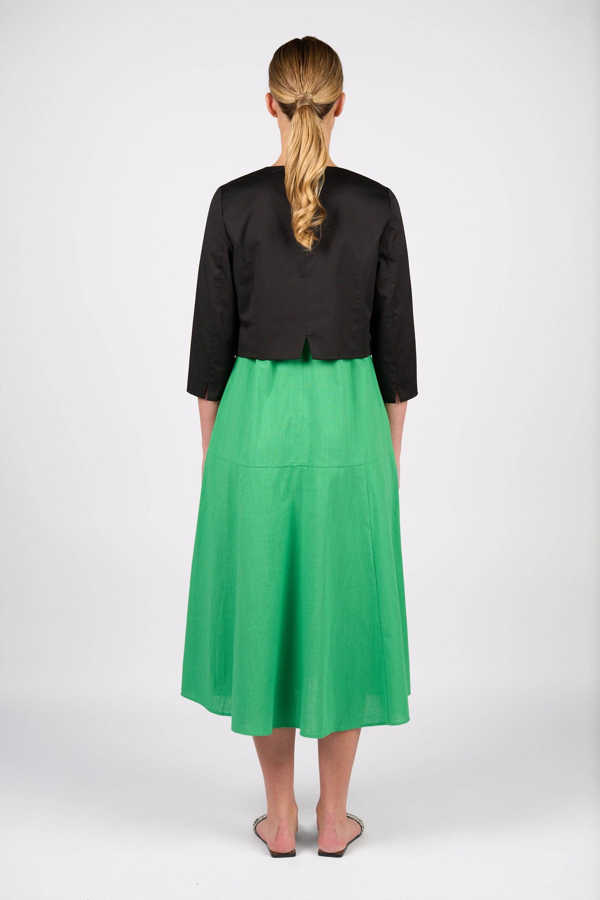 A woman with blonde hair in a low ponytail, seen from behind against a white background, wears a Vassalli Cropped Jacket - Black with 3/4 sleeves, a bright green midi skirt, and pointed flats.